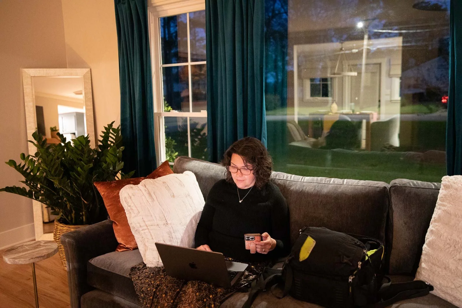 Woman with glasses sitting on a gray couch, using a laptop and holding a phone in a living room with large window and blue curtains at dusk.