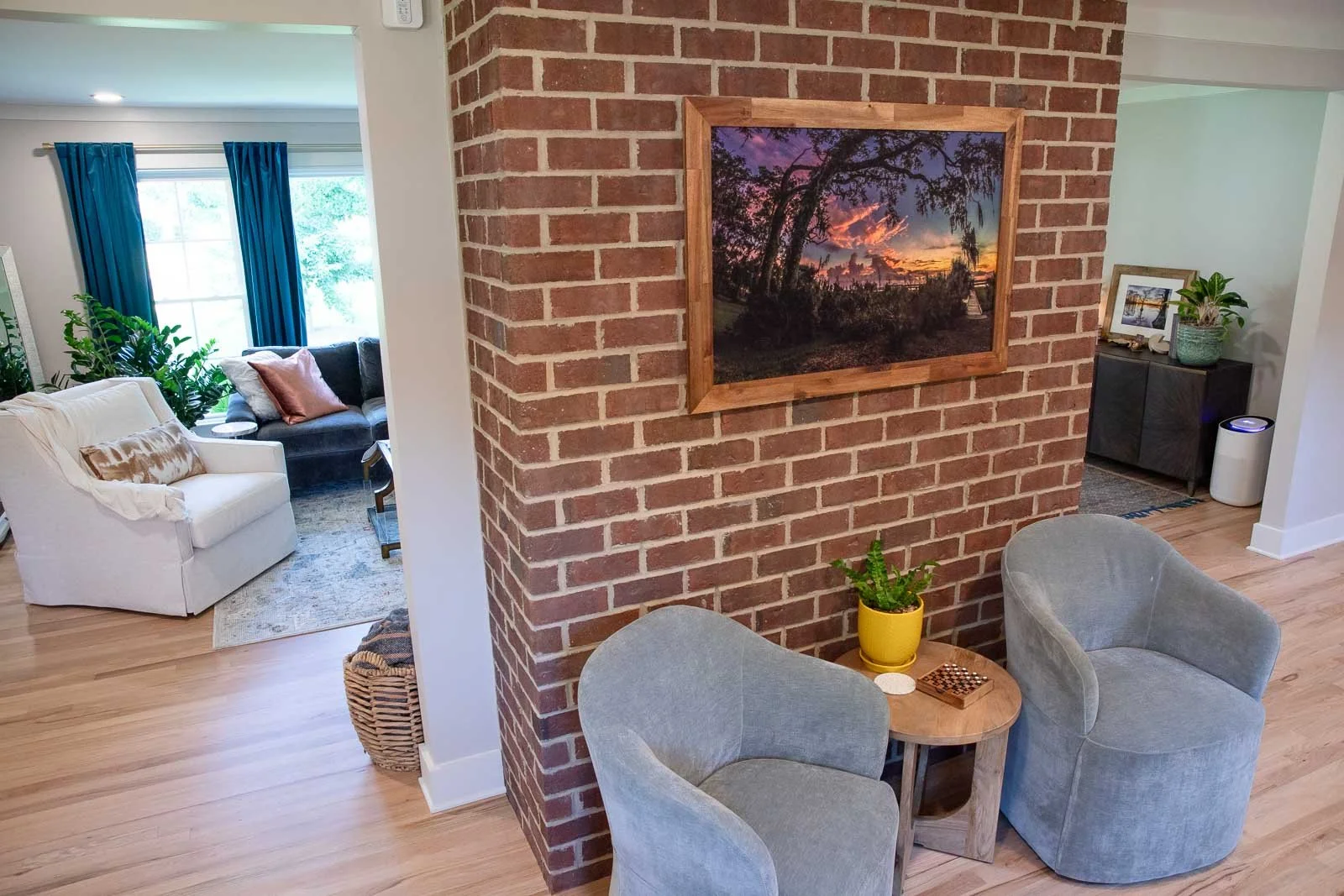 Interior view of a living room with a brick wall, two light gray armchairs, a small wooden table, a yellow potted plant, and framed pictures on the wall. In the background, there is a white sofa, a dark gray sofa with pink cushions, and large windows