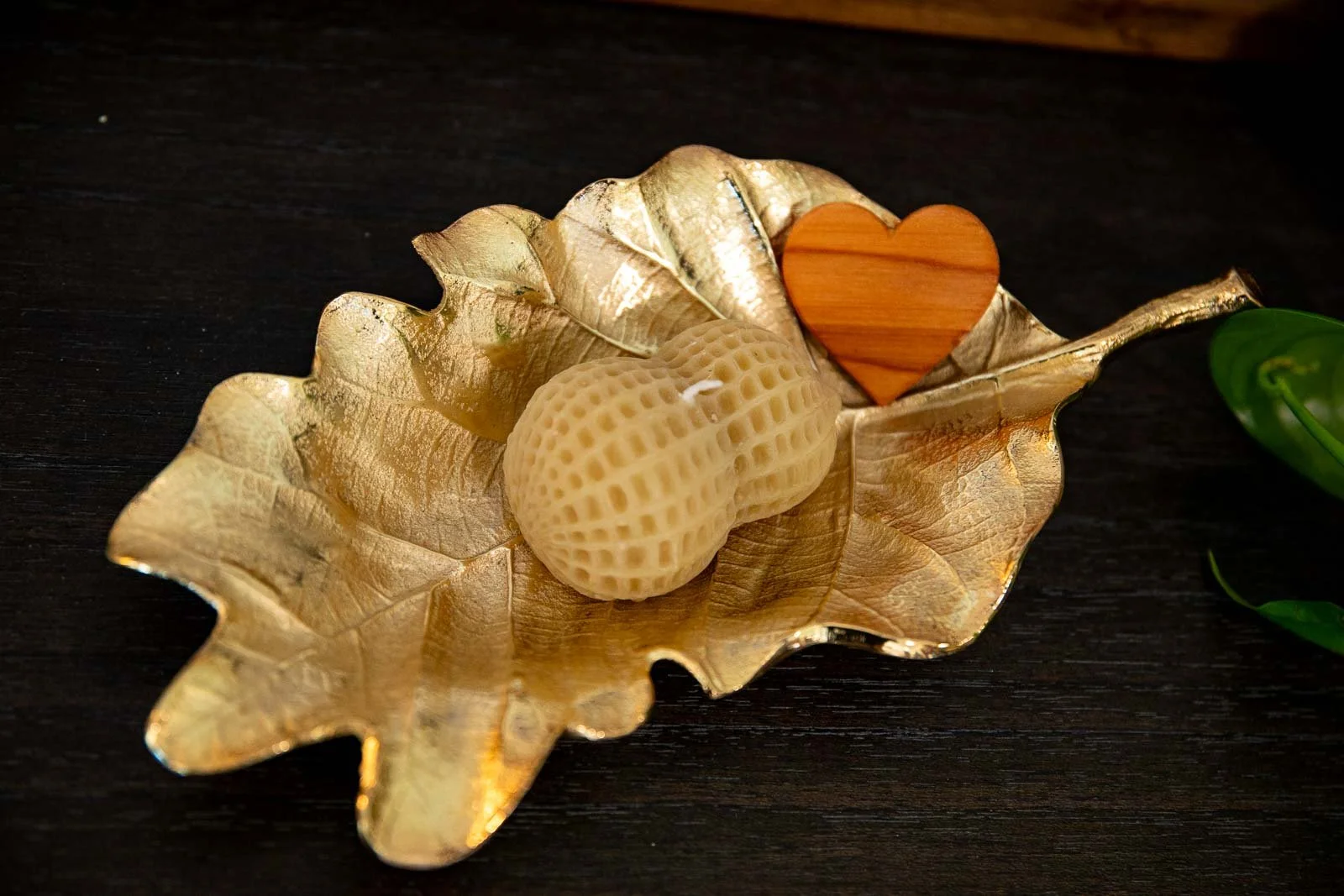 Decorative gold-leaf shaped dish holding a beige seashell and a small wooden heart, placed on a dark surface with part of a green plant visible on the right.