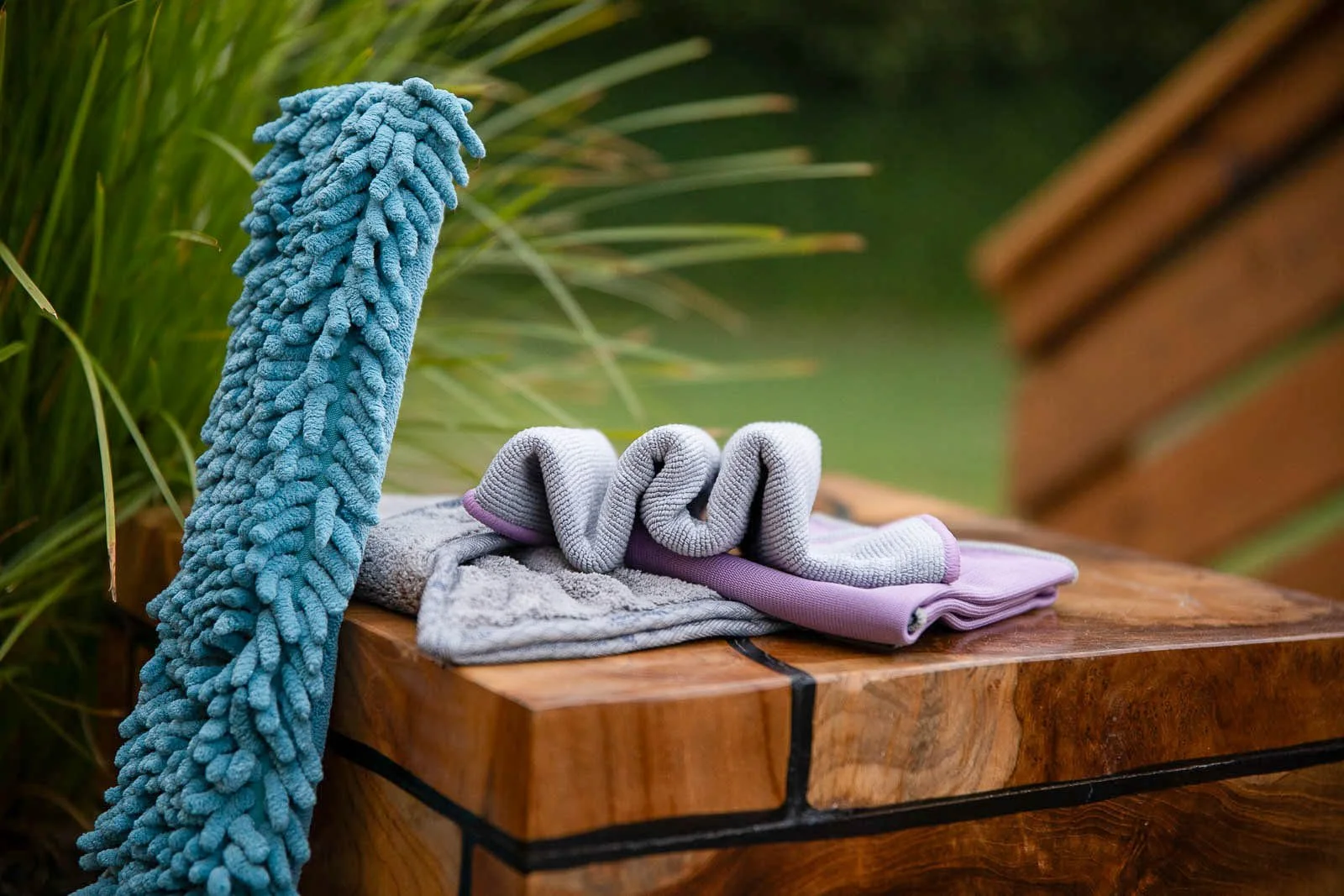 A rolled-up gray towel, a gray washcloth, and a purple yoga mat are on a wooden surface outdoors, with a blue towel hanging over the side and green foliage in the background.