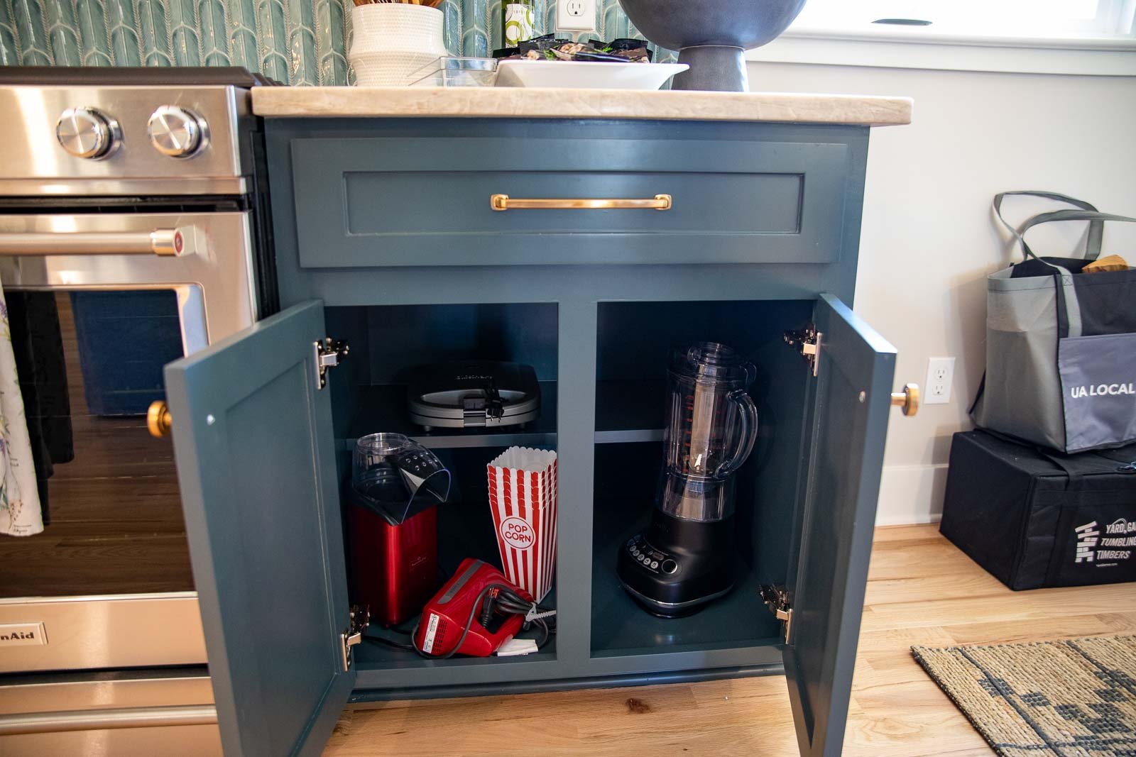 Open kitchen cabinet with kitchen appliances inside, including a blender, a popcorn maker, and a red handheld vacuum cleaner. The cabinet is blue with a drawer above and a beige countertop. To the left is a stainless steel stove, and to the right, pa