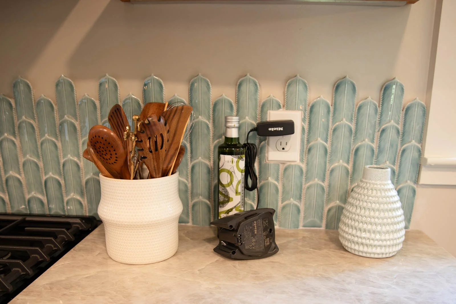 Kitchen countertop with a white textured utensil holder containing wooden and metal utensils, a small black electronic device, a decorative white ceramic vase, and a wall outlet with a black device plugged in. The background features a blue-green til