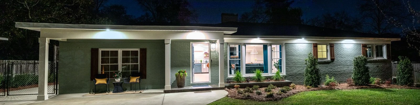 Exterior view of a house at night with its lights on, showing a front porch with chairs, a potted plant, and windows revealing a lit interior, surrounded by a lawn and shrubbery.