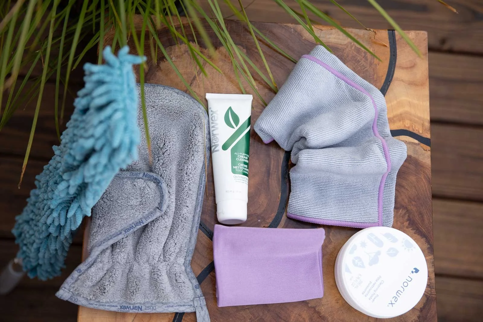 A wooden table with cleaning supplies, including gray cleaning gloves, a tube of natural all-purpose cream cleaner, a folded gray cleaning cloth, a lavender cleaning cloth, a disposable microfiber cleaning glove, and a round container of RenuLux clea