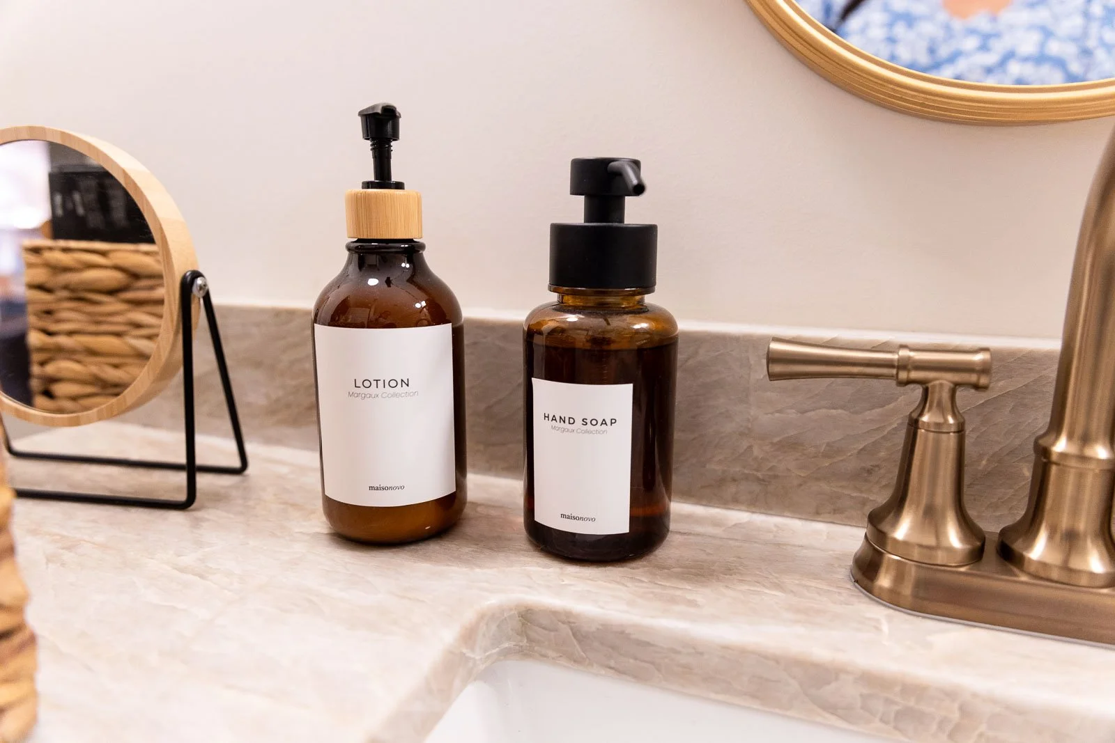 Brown bottles of lotion and hand soap with white labels on a bathroom countertop near a brass faucet and a round mirror.