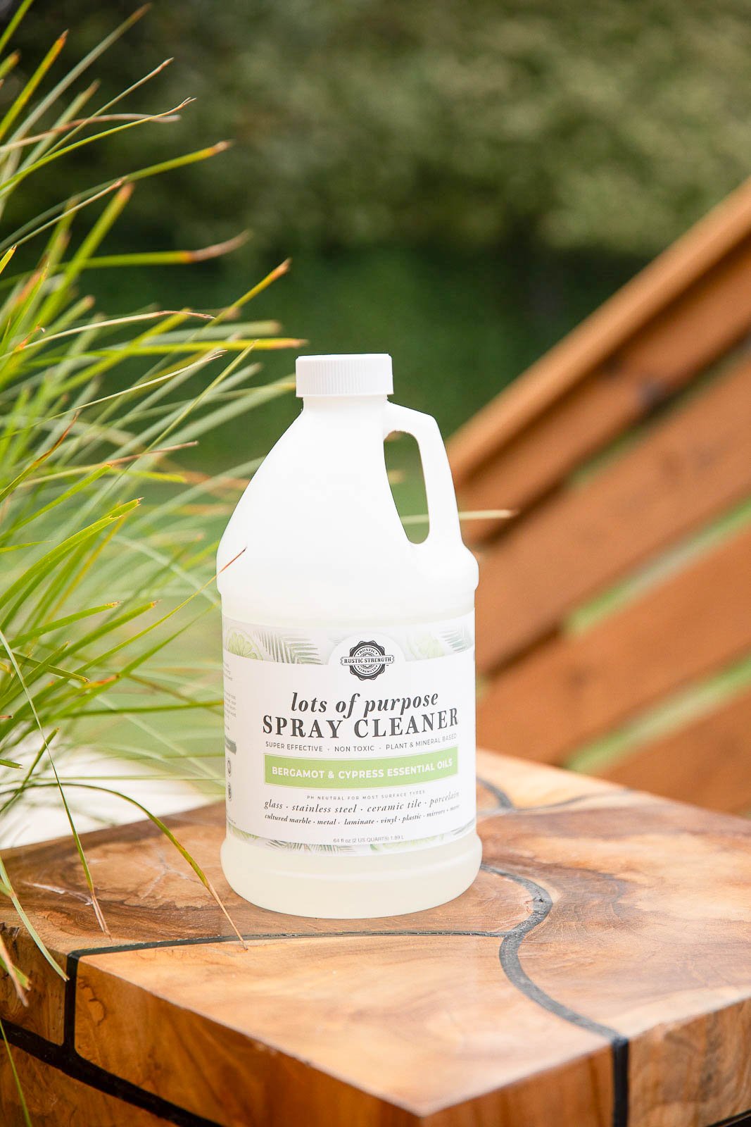 A bottle of spray cleaner labeled "Lots of purpose spray cleaner" with bergamot and cypress essential oils, placed on a wooden surface outdoors with greenery and a wooden fence in the background.