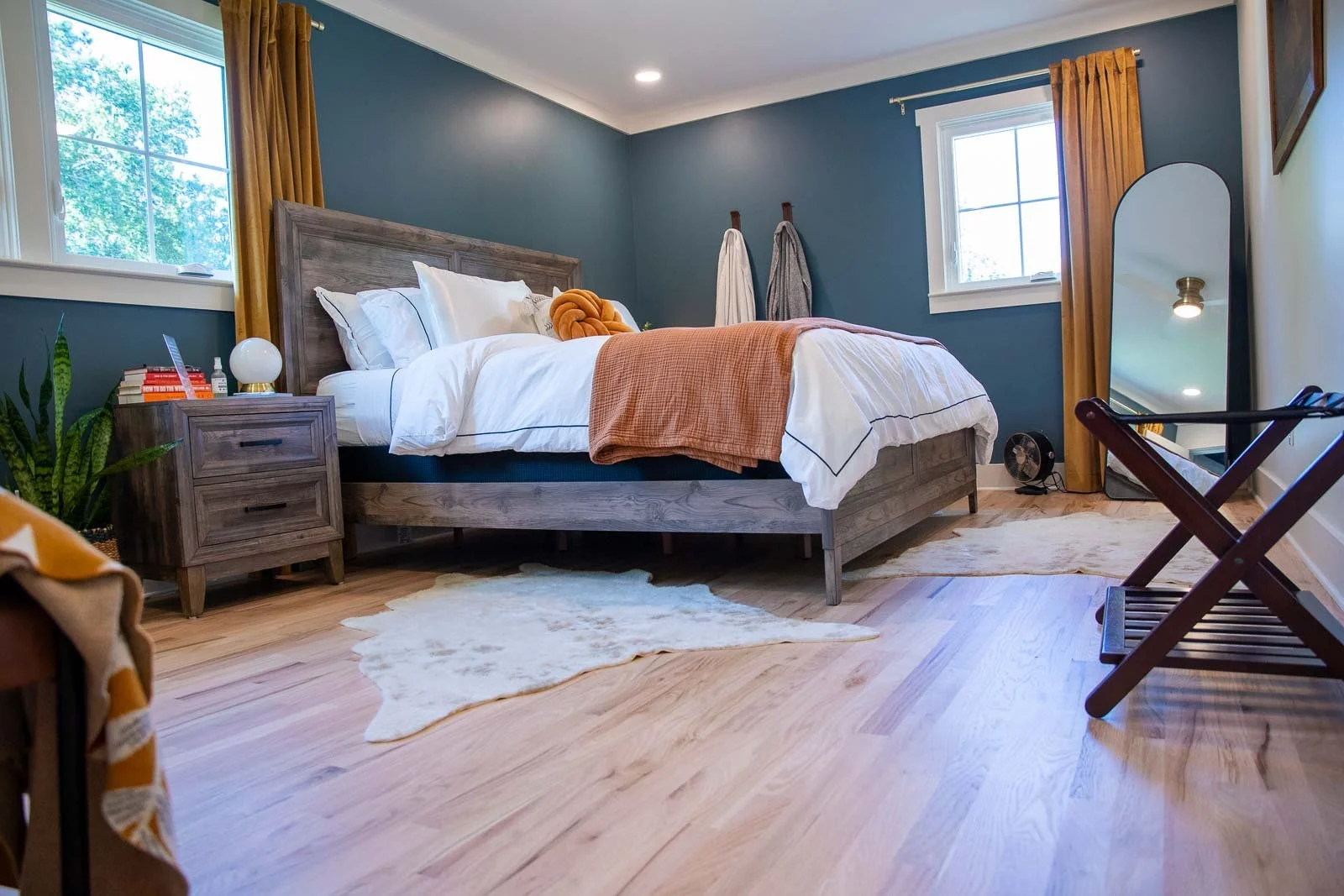 A bedroom with green walls, wooden furniture, a bed with white bedding, and yellow curtains. There is a mirror and a suitcase rack near the window, with a small fan on the floor.