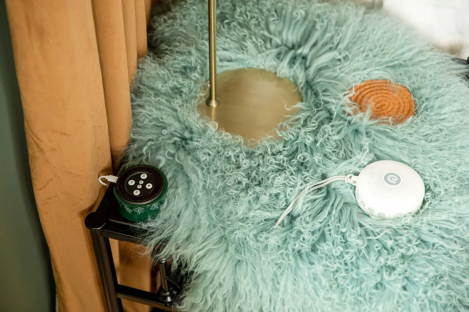 A bedside table with a retro green shaggy lamp, a portable speaker, and a white smart speaker on a plush mint green fuzzy bedspread next to mustard-colored curtains.
