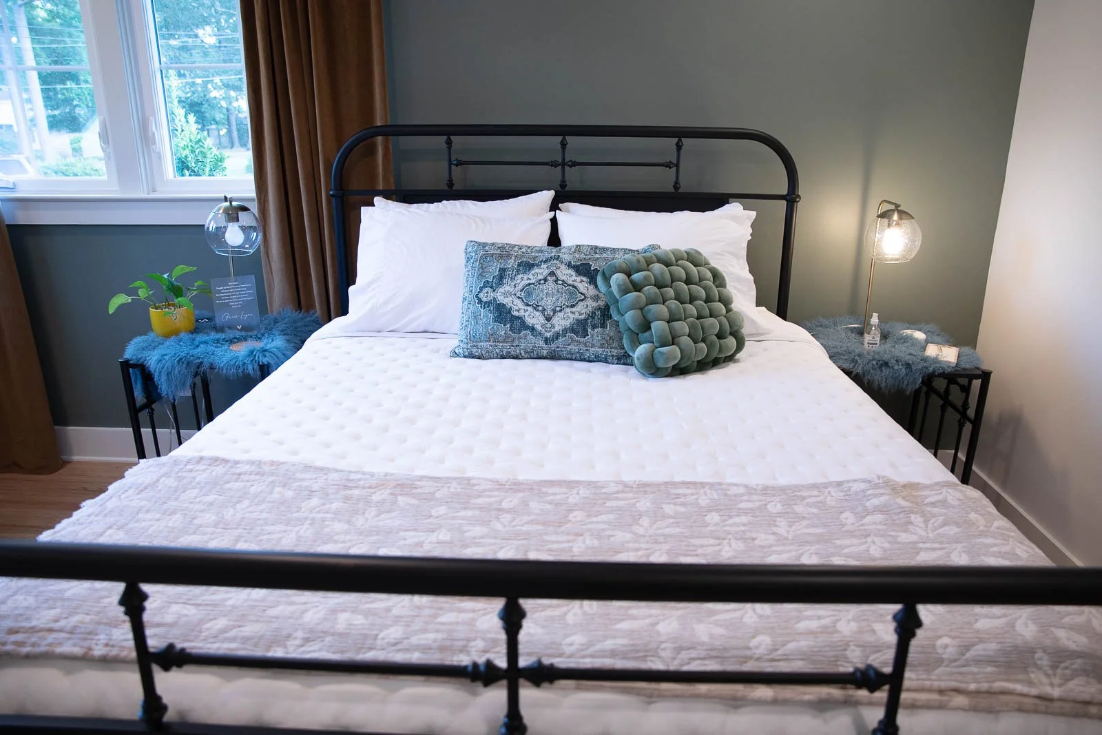 A neatly made bed with white bedding, a decorative pillow, and a plush green pillow with a bubble texture. The bed has a black metal headboard and is flanked by two bedside tables with lamps, one with a glass dome cover and the other with a furry clo