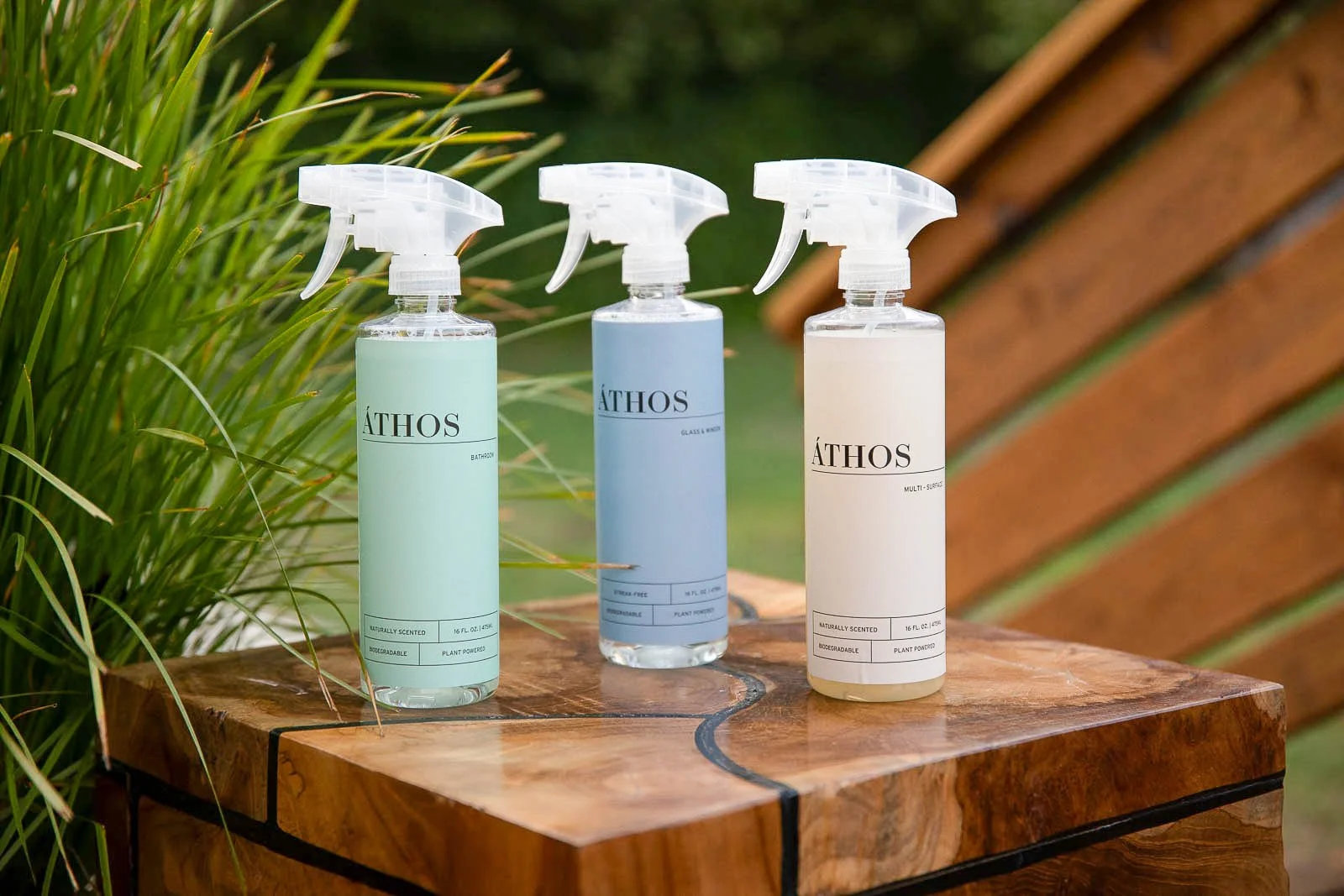 Three spray bottles of cleaning products labeled 'ÁTHOS' placed on a wooden surface outdoors, surrounded by greenery and a wooden fence.