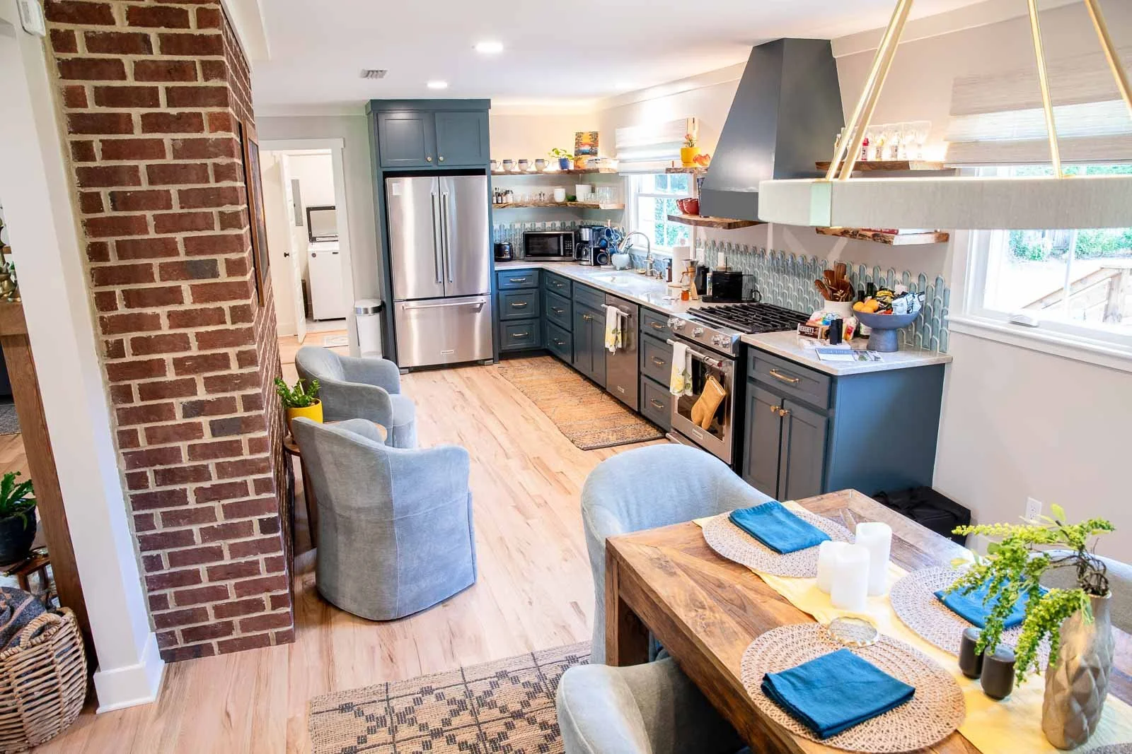 A modern kitchen with blue cabinets, stainless steel appliances, open shelving, and a dining area with a wooden table and blue napkins.