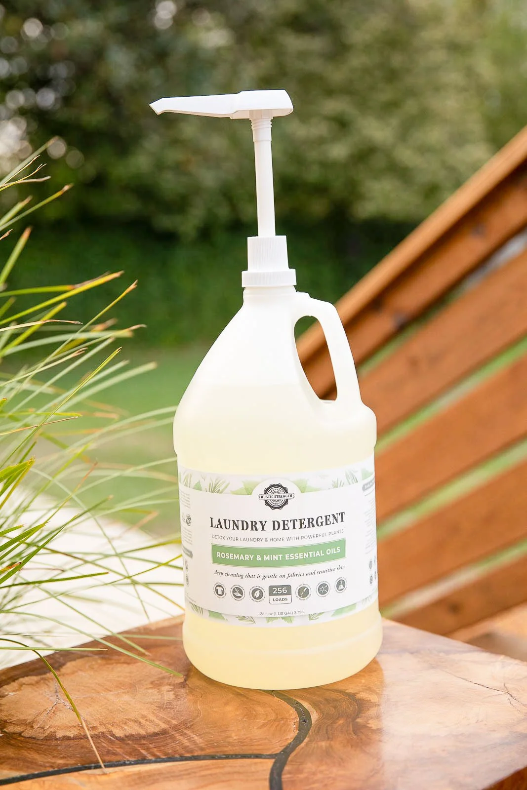 White plastic bottle of laundry detergent with pump dispenser on a wooden table outdoors, with grass and trees in the background.