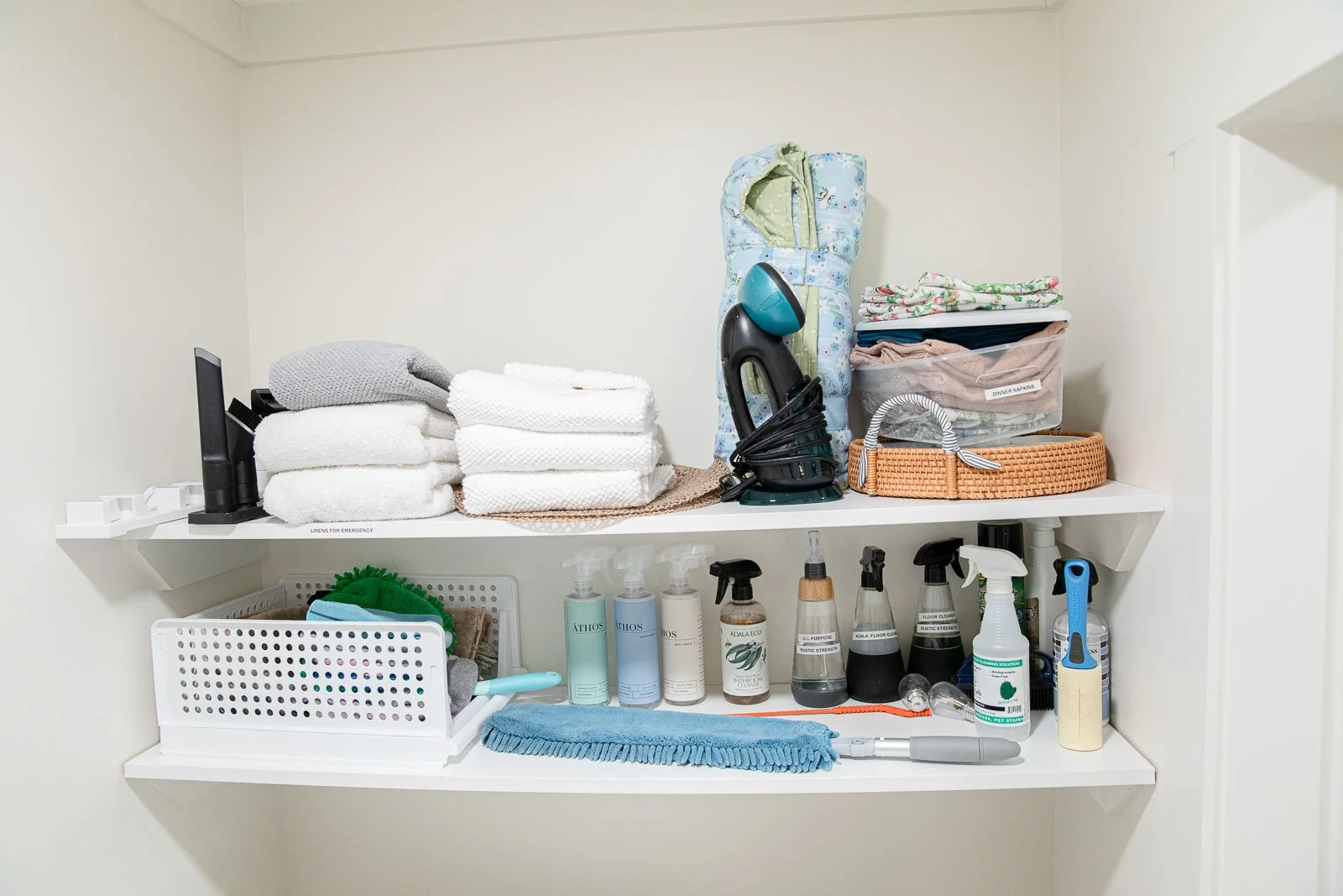 Two white shelves stocked with cleaning supplies, towels, cloths, and miscellaneous household items