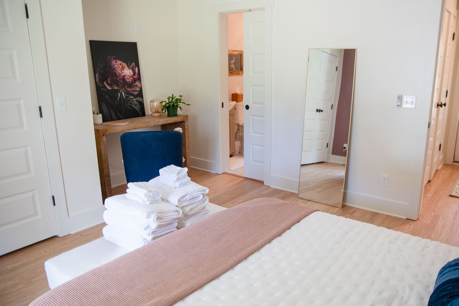 A bedroom with a bed, stack of white towels on the bed, a blue chair, a wooden desk with a potted plant and artwork, a large mirror leaning against the wall, and an open door leading to a bathroom.