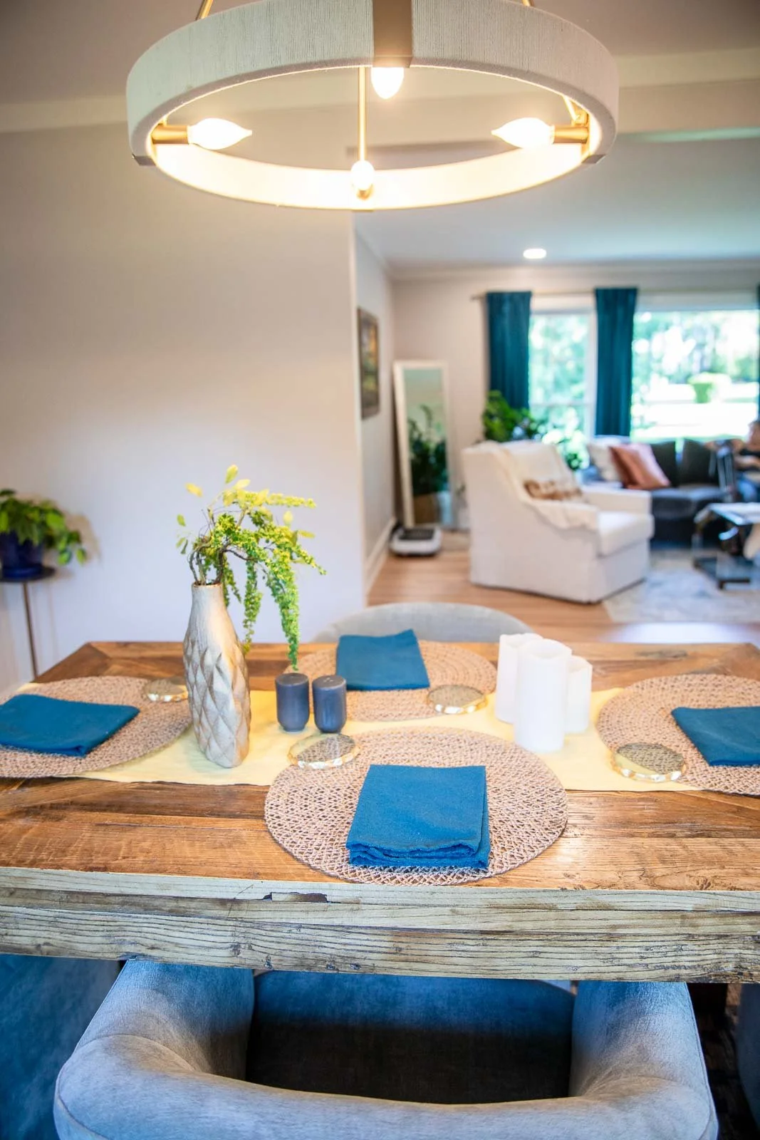 A dining table set for four with blue napkins, woven placemats, and a decorative vase with greenery, in a well-lit living room and dining area.