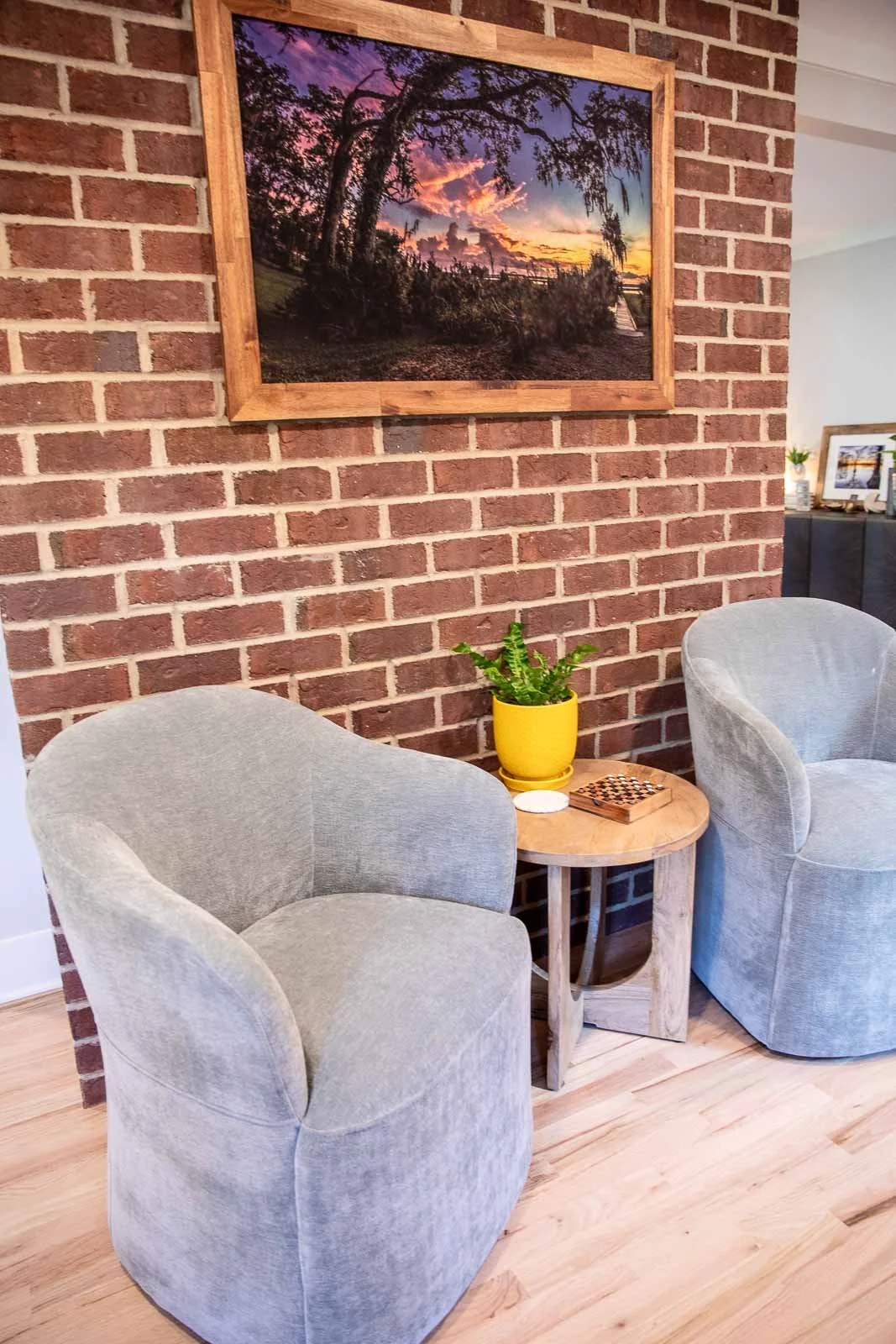 A cozy living room corner with two light gray armchairs and a small round wooden table, featuring a yellow potted plant, a chessboard, against a brick wall with a framed sunset landscape photograph above.
