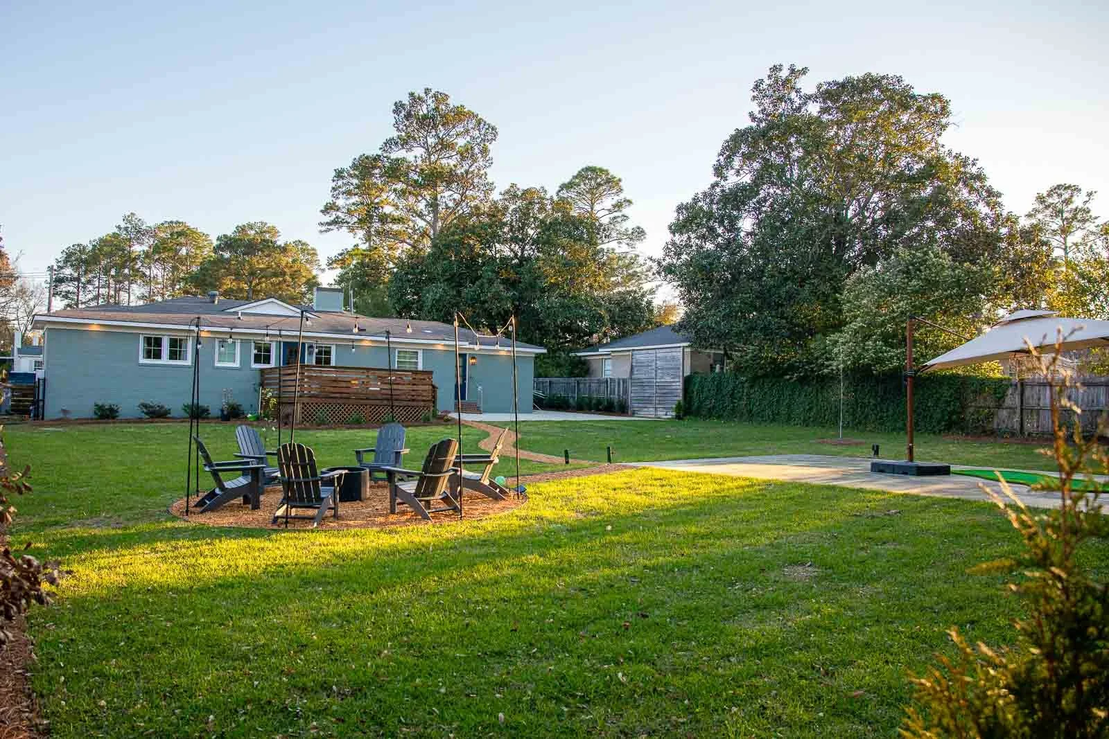 Backyard with Adirondack chairs around a fire pit, string lights hanging, grassy area, trees, a house in the background, and a patio with a white umbrella.