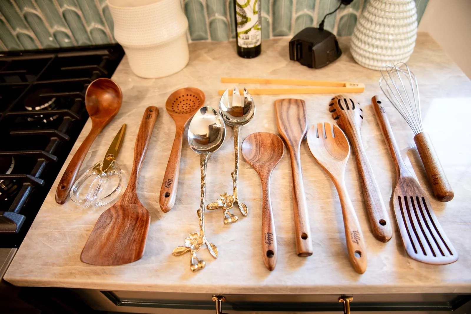 A collection of various kitchen utensils, including wooden spoons, metal spoons, a whisk, tongs, and spatulas, arranged on a light-colored countertop.