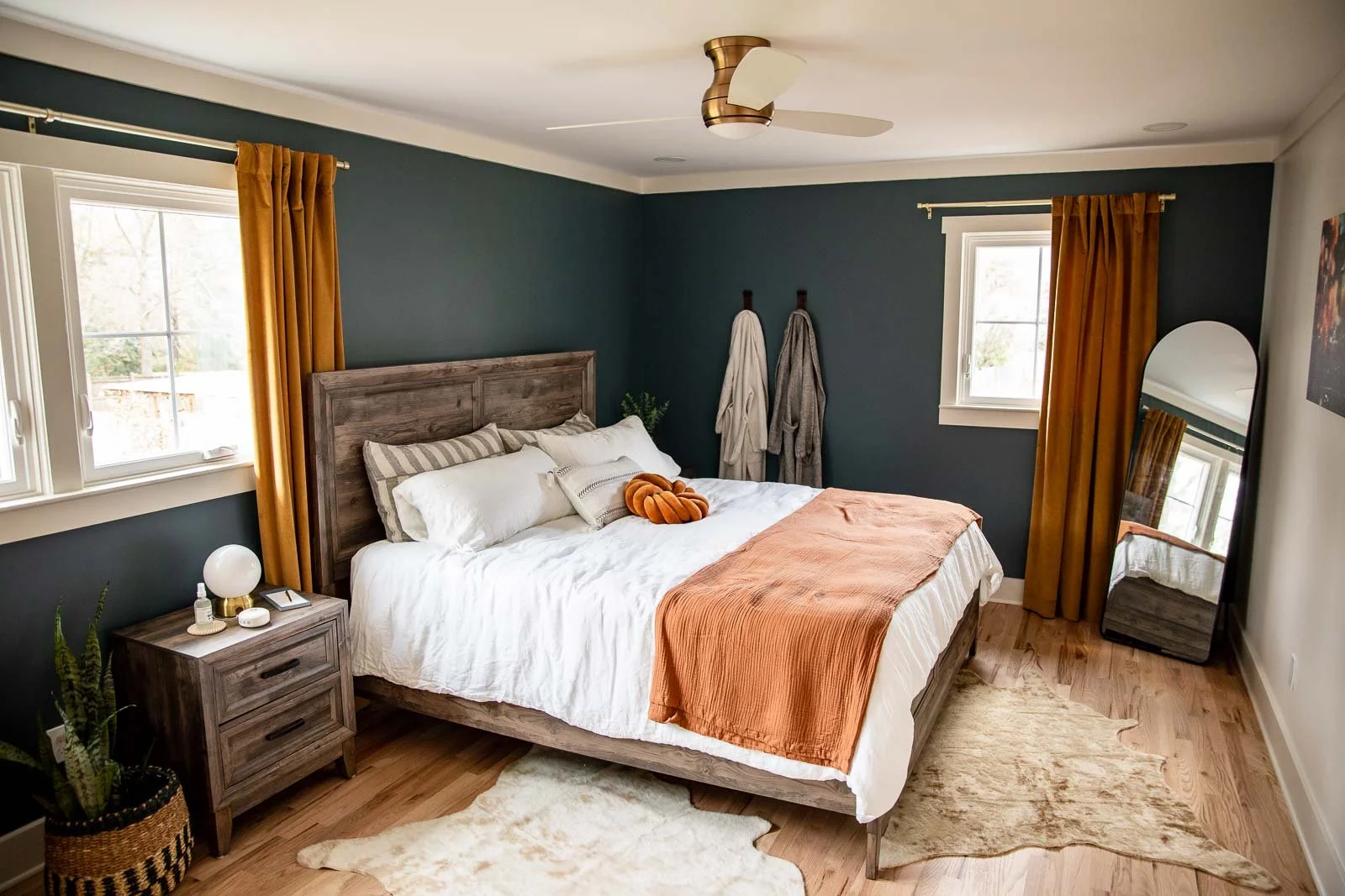 A cozy bedroom with dark teal walls, a wooden bed frame with white bedding and a rust-colored throw blanket. Two windows have mustard yellow curtains, and there is a mirror leaning against the wall. A nightstand with a round lamp and items is next to