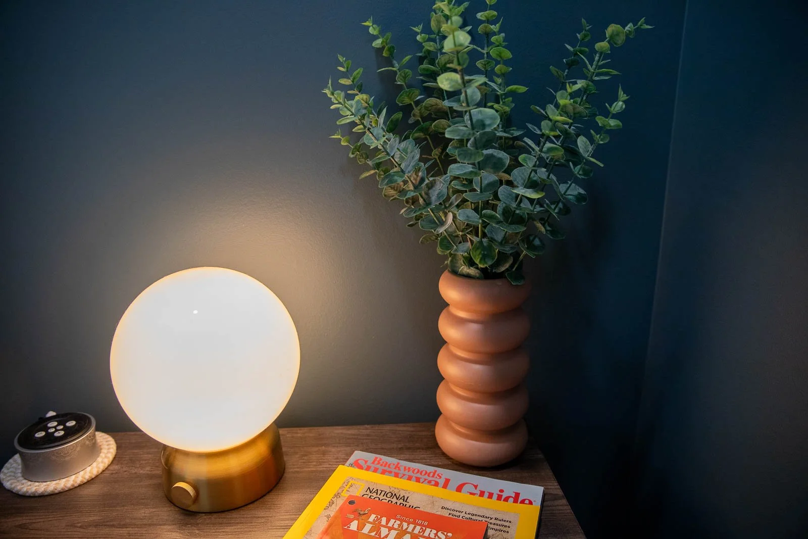 A glowing round table lamp with a gold base, a small remote control on a white braided coaster, a stack of magazines, and a tall green plant in a tan ribbed vase on a wooden surface against a dark wall.