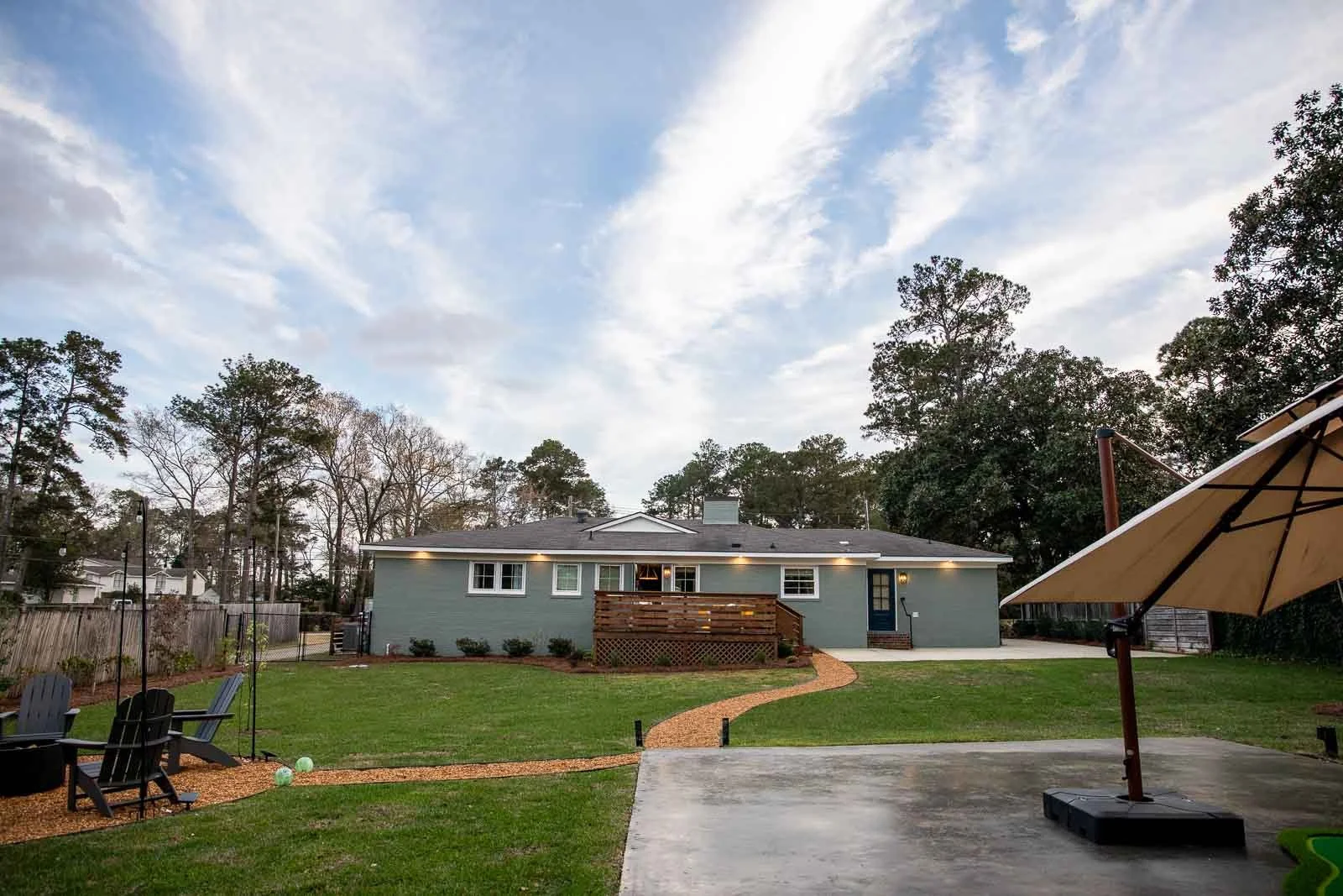 A suburban backyard with a green lawn, potted plants, outdoor chairs, a large patio umbrella, and a blue house with a wooden deck, set against a sky with clouds and tall trees in the background.