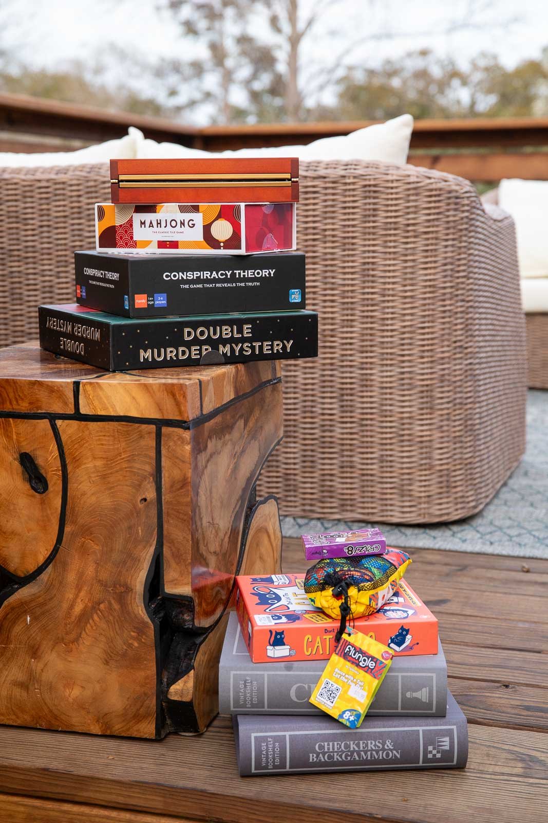 Stacked board games on and beside a wooden table outdoors, including Mahjong, Conspiracy Theory, Double Murder Mystery, and Checkers & Backgammon, with a small colorful drawstring bag and a card game on top.