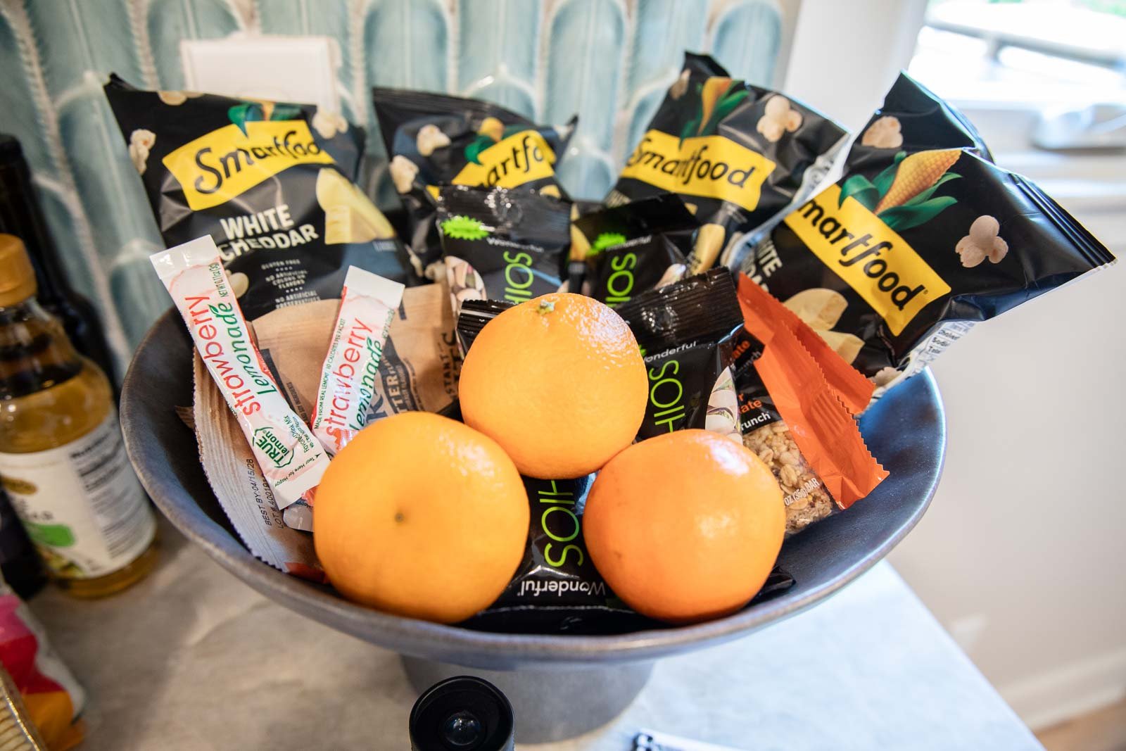 A gray bowl containing three fresh oranges, several packets of Smartfood white cheddar popcorn, and packets of Singanios taco sauce, granola, and an orange-wrapped snack, placed on a kitchen counter.
