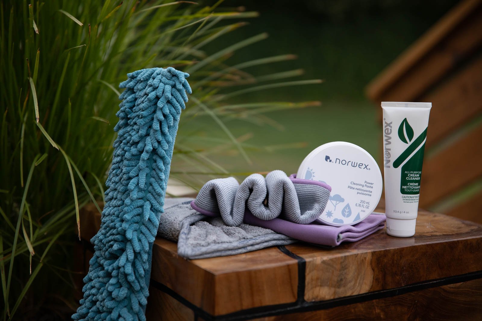 Outdoor wooden table with cleaning and grooming supplies, including a blue microfiber cloth, gray cleaning cloth, purple and gray socks, Norwex cleaning paste in a white container, and Norwex cream cleaner tube, with green foliage in the background.