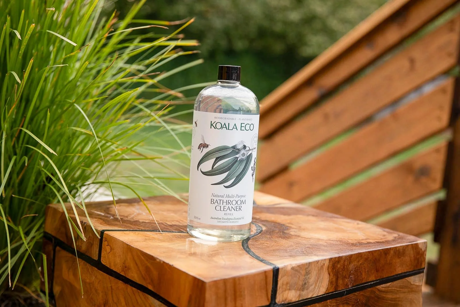 A bottle of Koala Eco bathroom cleaner with eucalyptus illustration on the label, placed on a wooden table outdoors with green plants and a wooden bench in the background.