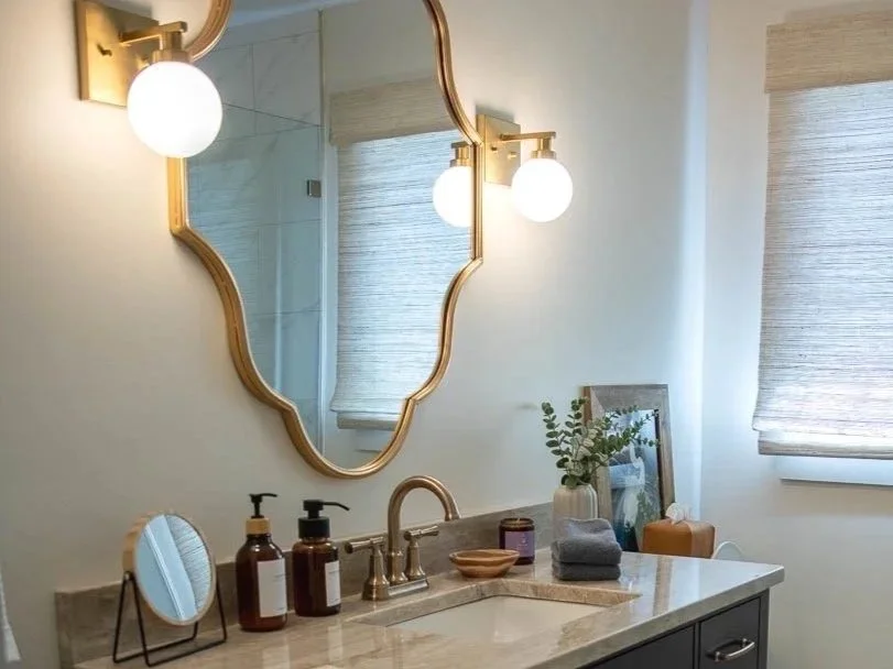 Bathroom with a dark vanity, beige marble countertop, and gold fixtures, including a mirror and light fixtures. The bathroom has a window with a beige textured shade, a plant, and toiletries on the counter.