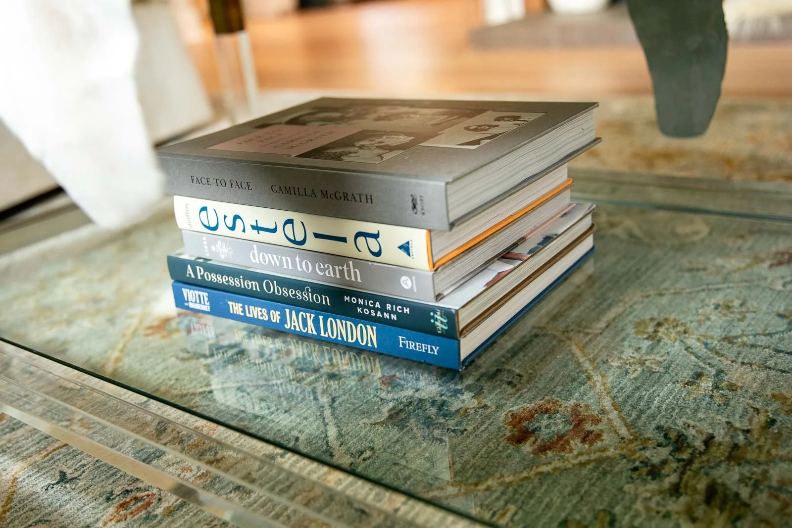 A stack of five books on a glass table, with a patterned rug underneath and part of a chair and a potted plant in the background.