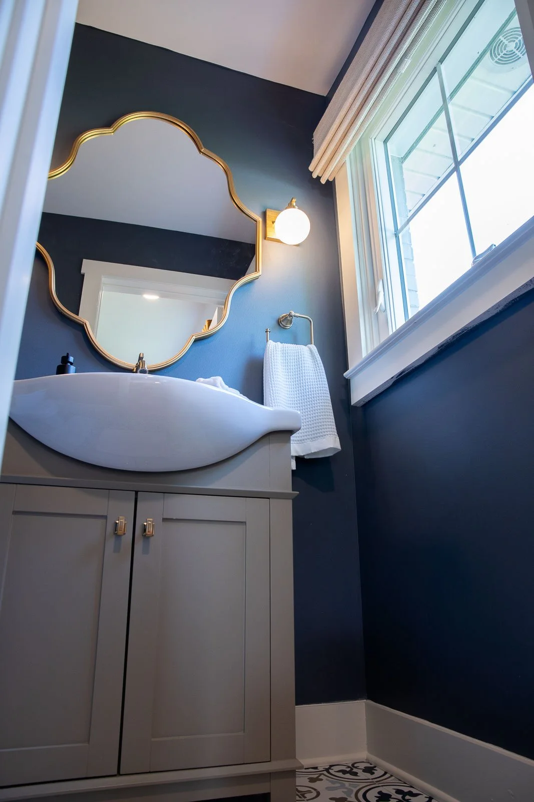 A bathroom with a gray vanity, white sink, decorative mirror, white towel, wall light, and a window with cream curtains.