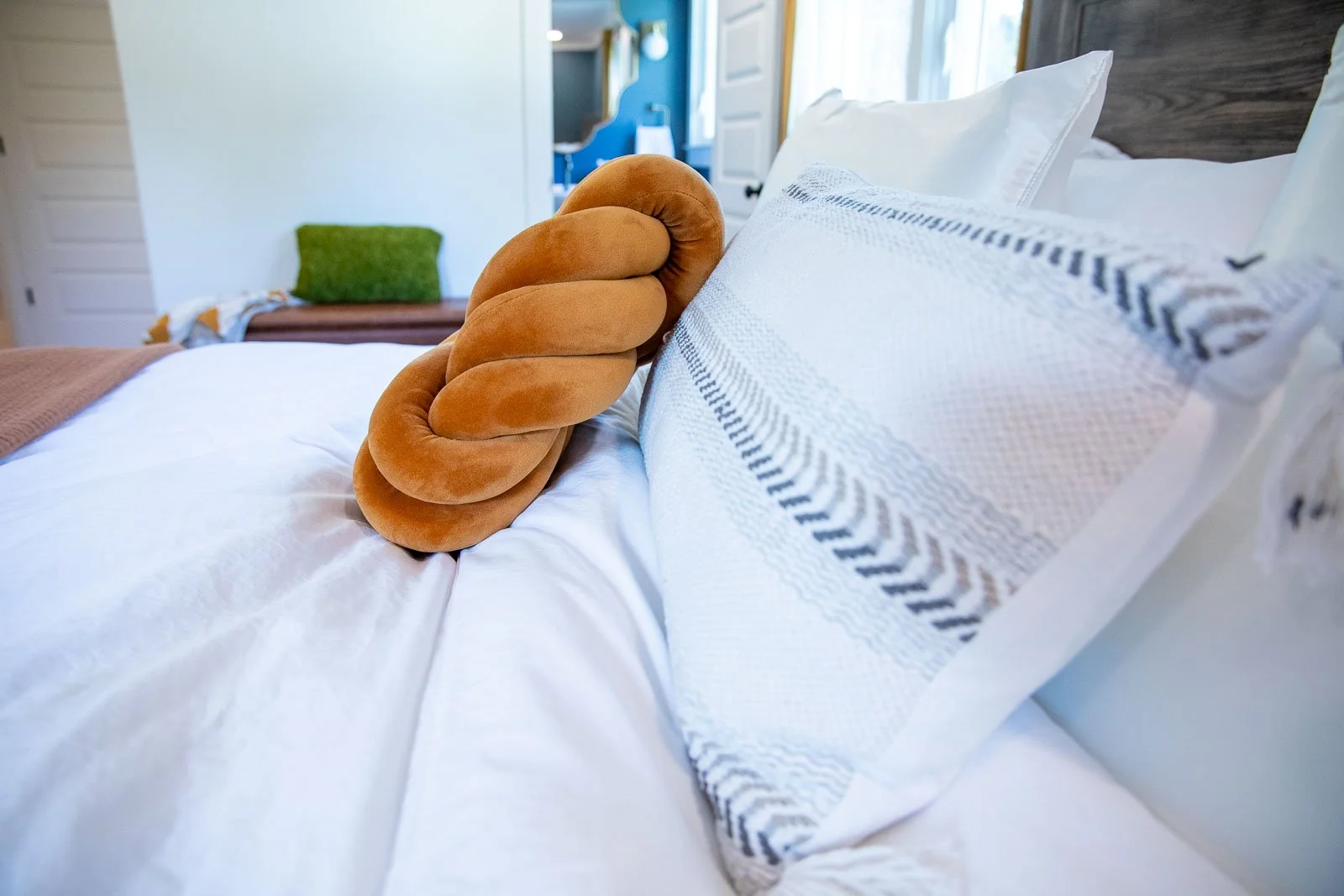 A bed with white linens and pillows, a large plush twisted brown pillow, and a wooden headboard in a cozy room with a bench and window in the background.