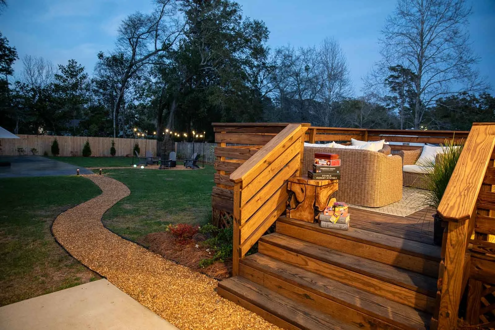 A backyard patio area with wooden stairs leading up to a seating area with wicker furniture, surrounded by a wooden fence, with trees in the background, and a winding gravel path leading to a lawn with chairs and string lights.
