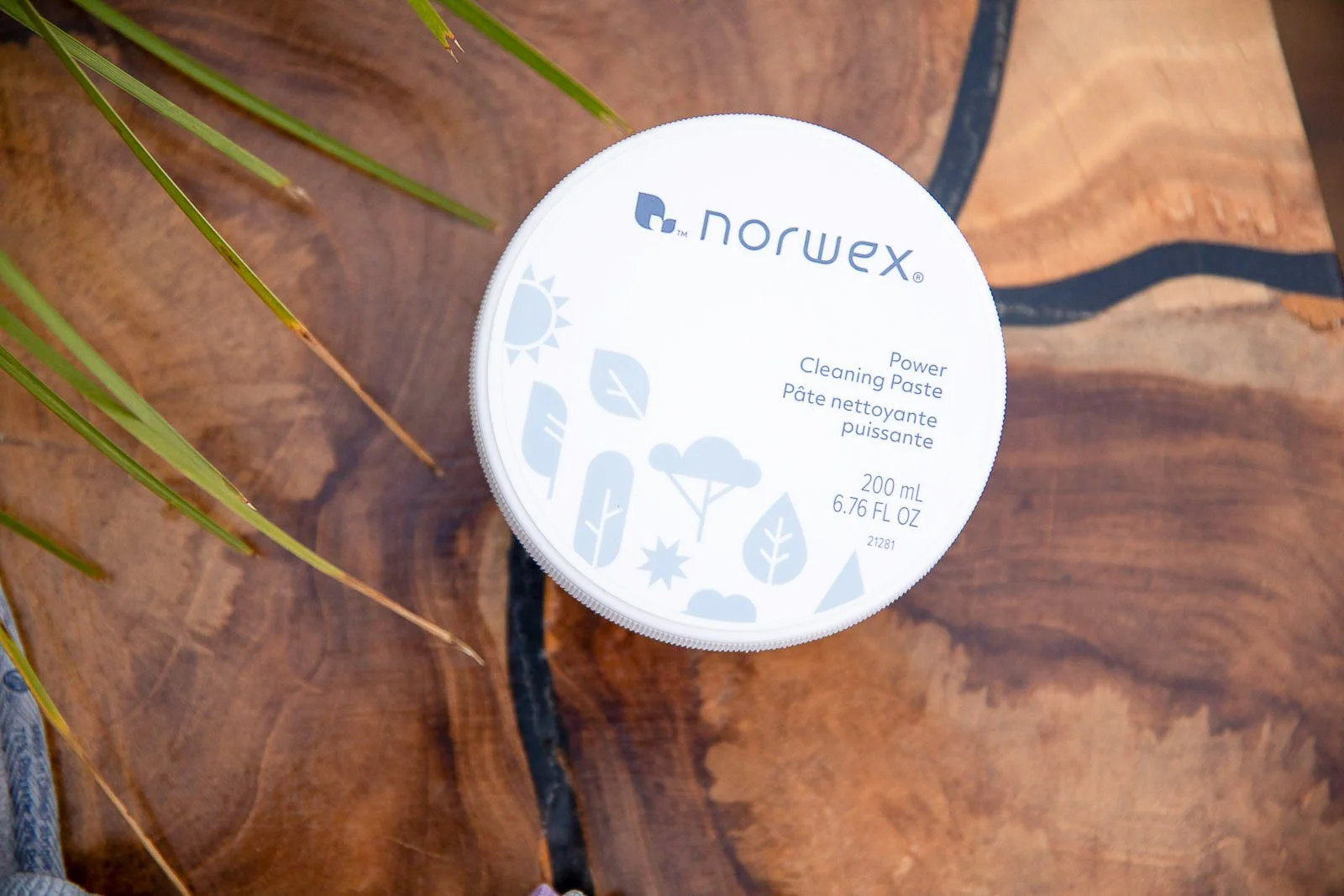 A round container of Norwex Power Cleaning Paste placed on a wooden surface with a plant leaf partially visible on the left side.