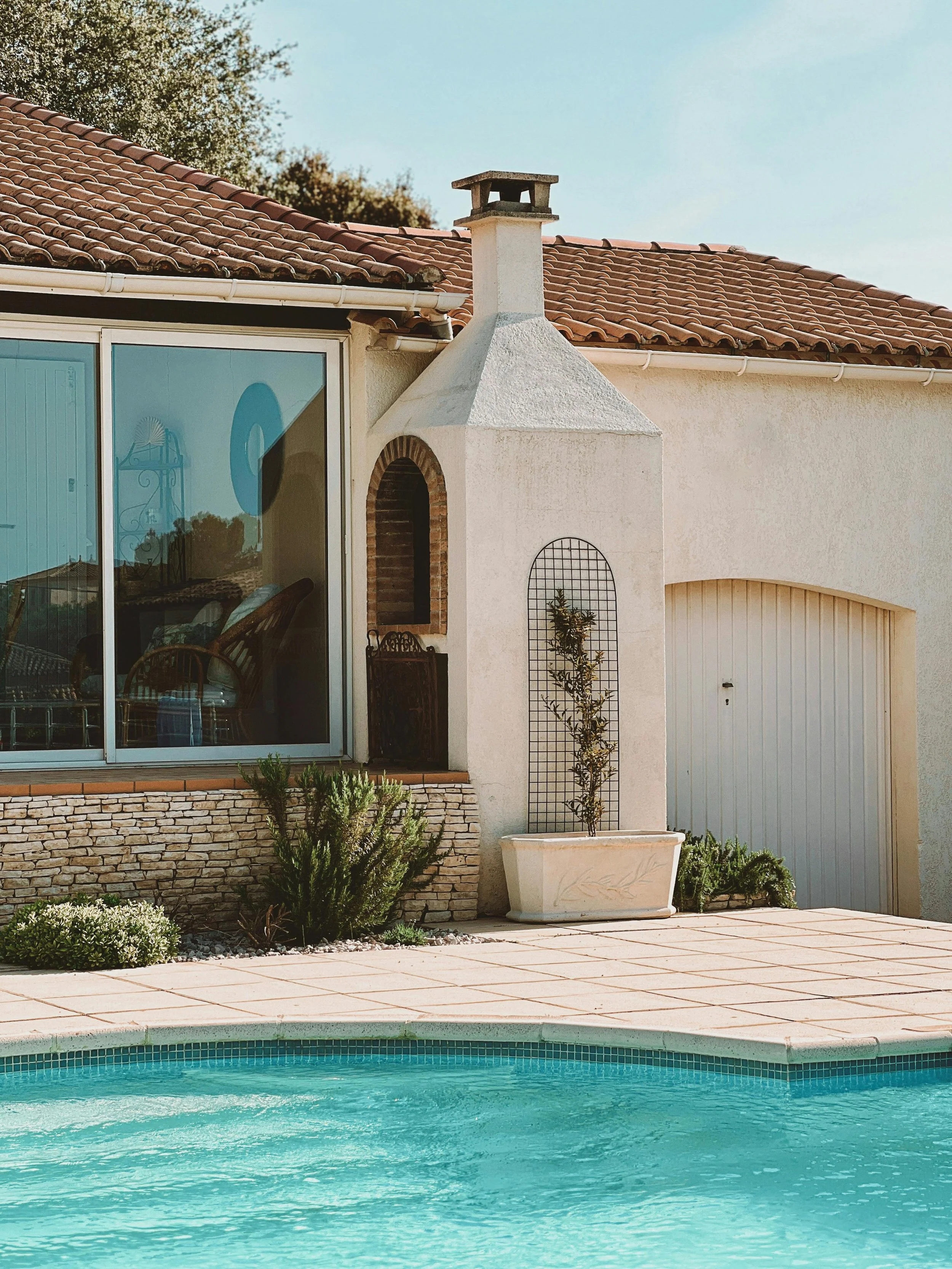 Side view of a house with a swimming pool in the foreground, featuring a chimney, large window, small door, garden plants, and a garage door under a clear sky.