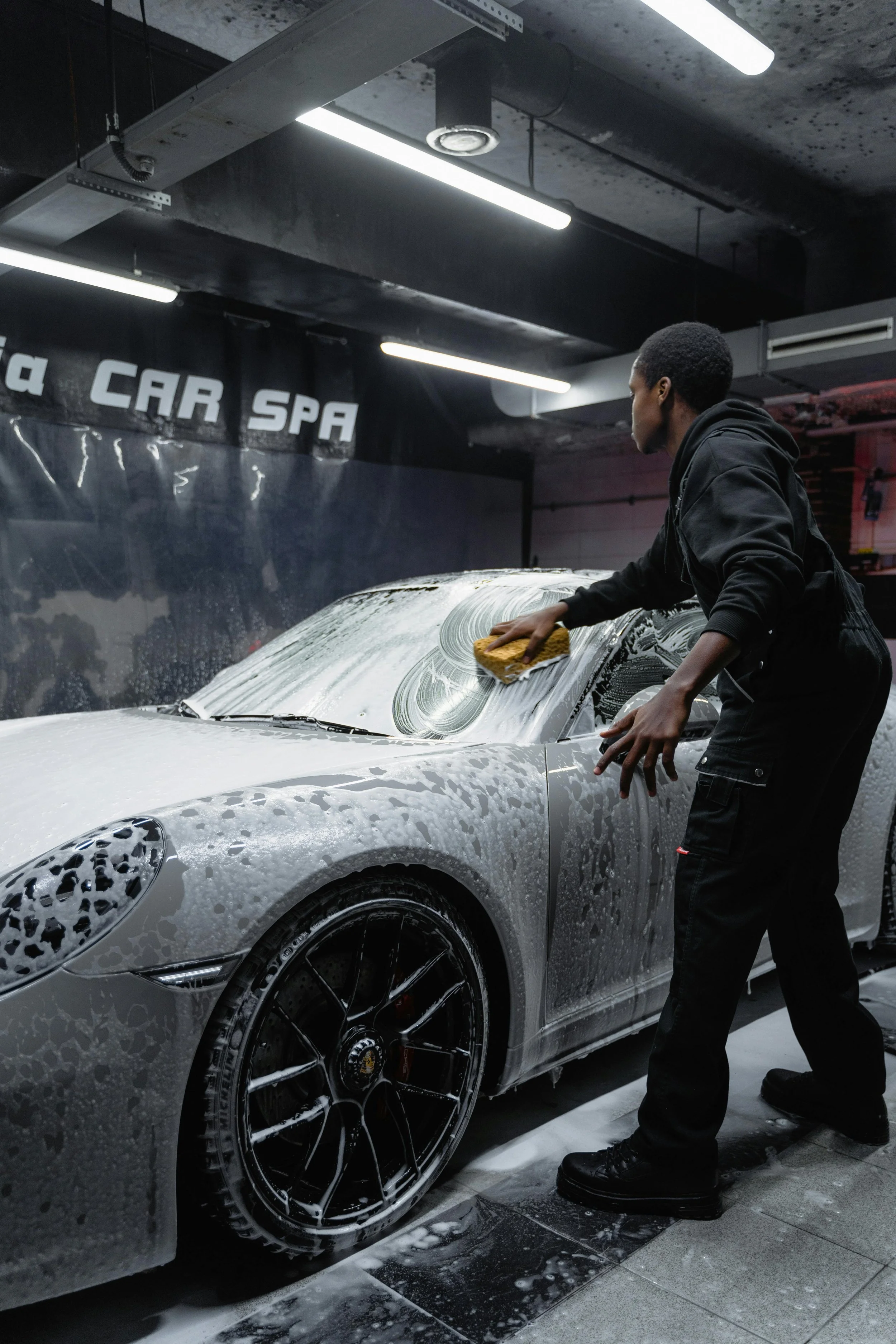 A person washing a silver sports car with a yellow sponge at a car wash or auto spa, with soap and foam on the vehicle.