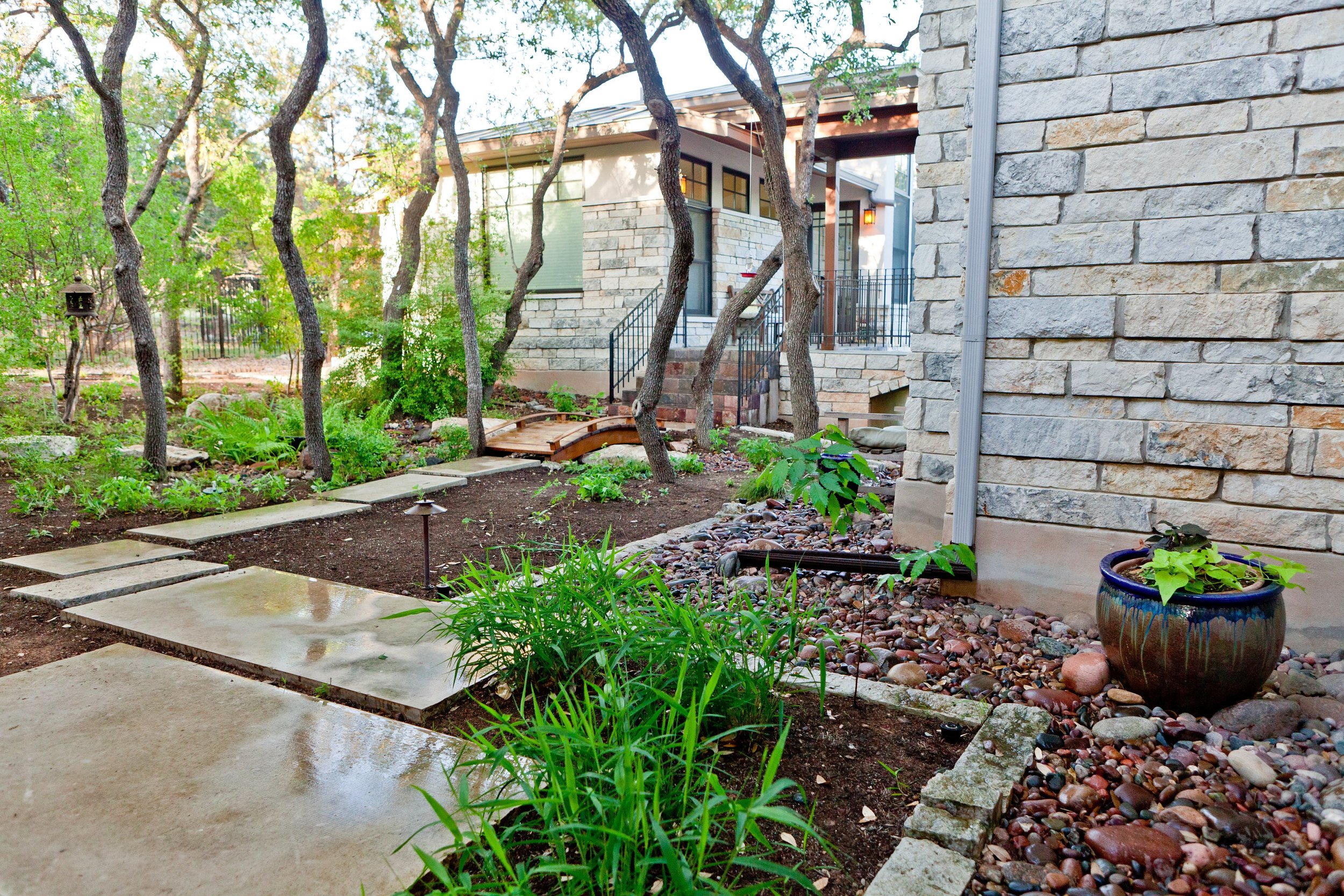 A backyard garden with a stone walkway, small trees, potted plants, and a house with stone walls and steps leading up to a porch.