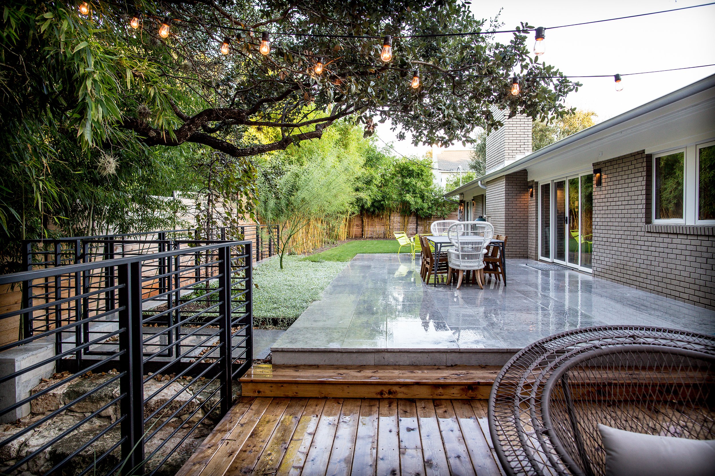 Backyard patio area with dining table and chairs, lush greenery, string lights overhead, and a wooden deck leading to the patio.
