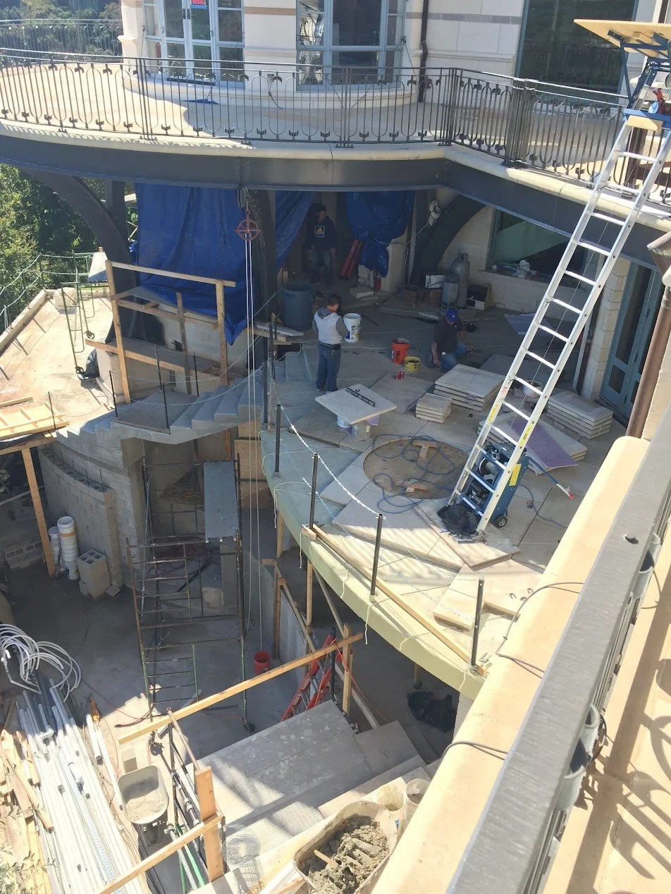 Construction site with workers installing a pool, cantilevered stone patio, ladders, scaffolding, and various construction materials.