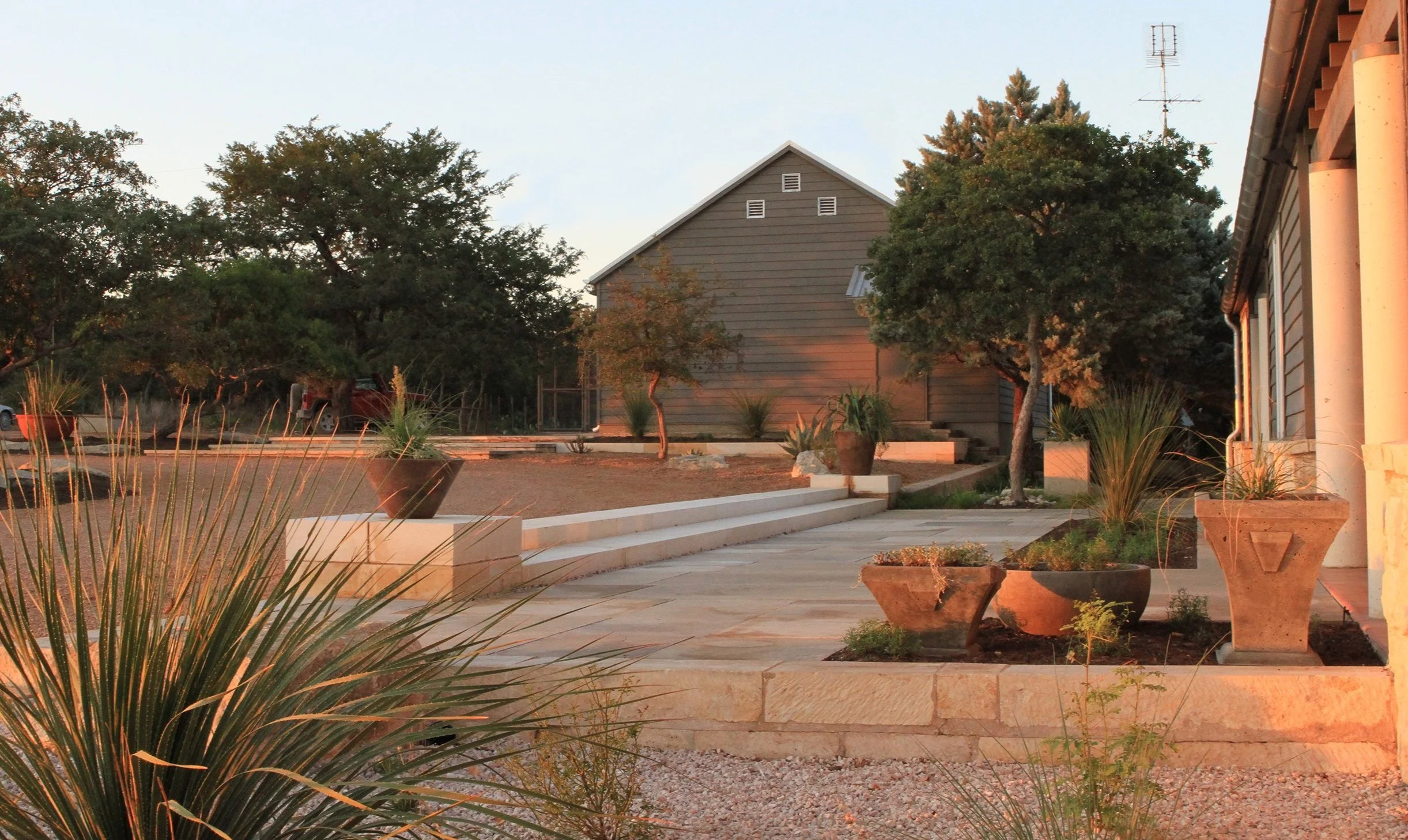 A landscaped backyard at sunset with potted plants, trees, and a wooden house in the background.