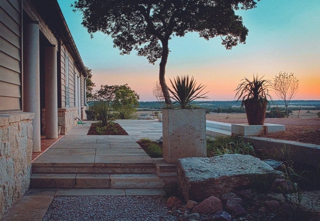 A modern outdoor patio with steps leading to an open landscape, featuring potted plants, trees, and a sunset sky.