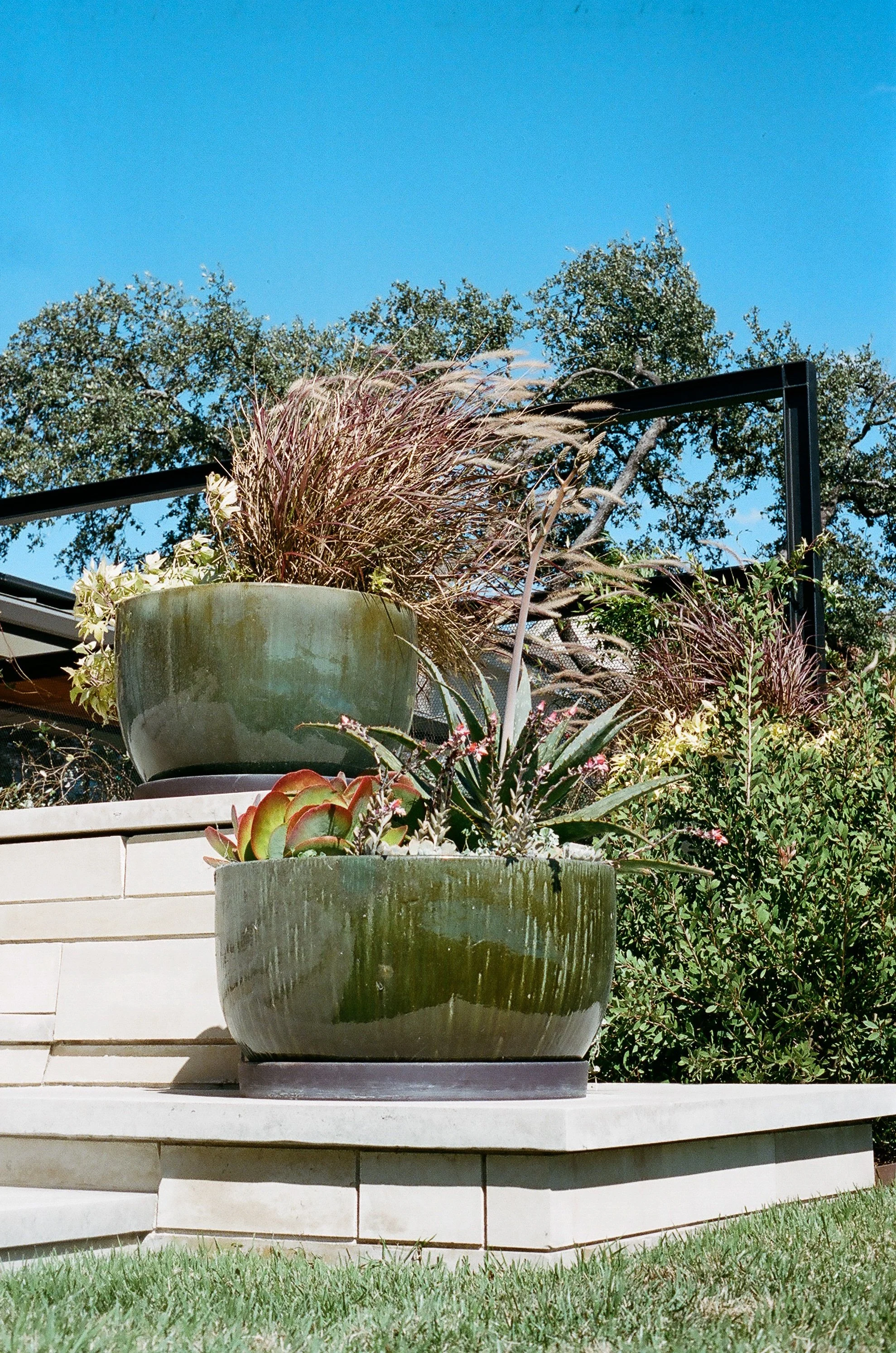 Two large green planters with various plants on a stone ledge outdoors under a blue sky.