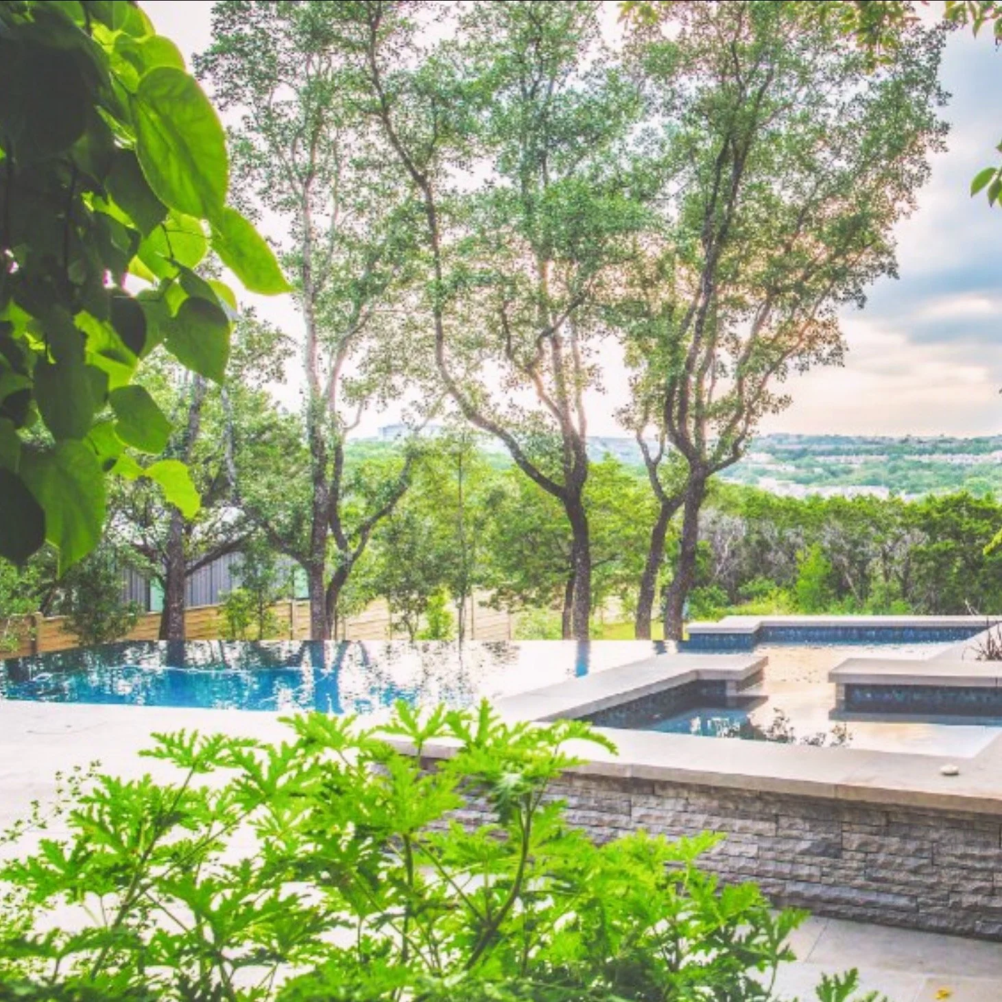 View of a swimming pool in a backyard with a stone ledge, surrounded by trees and green foliage, overlooking a cityscape in the distance on a partly cloudy day.