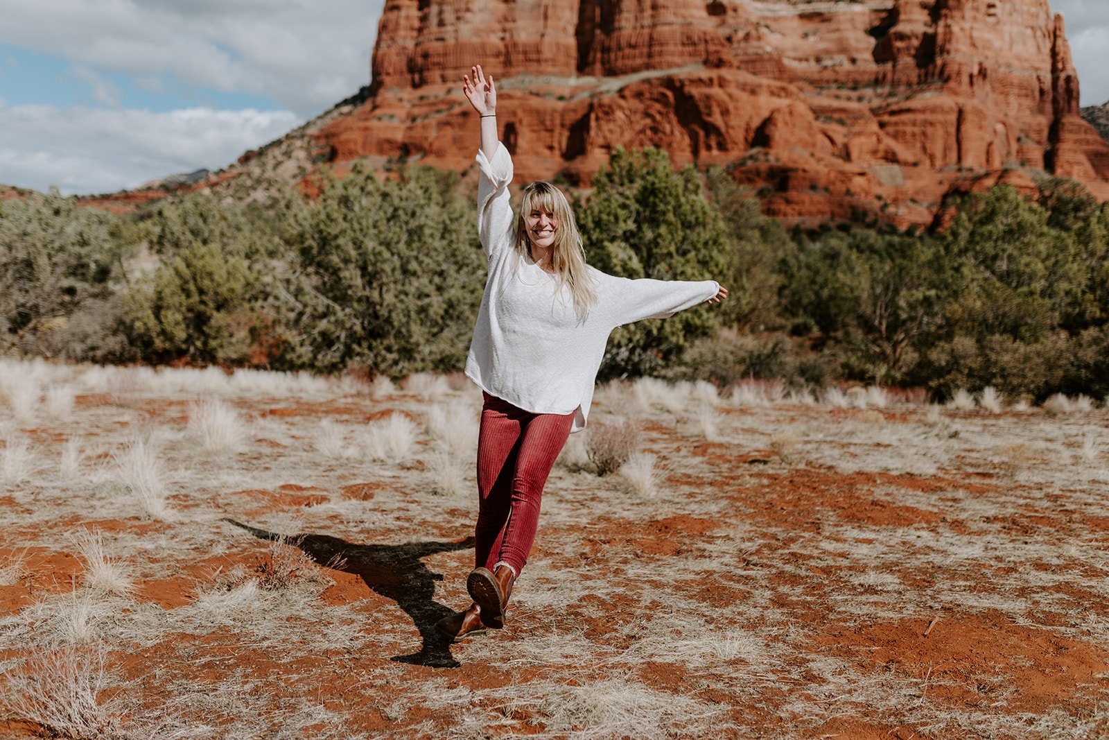 Woman in white sweater and red pants smiling with arms raised in a desert landscape with red rock formations and green shrubs in the background.