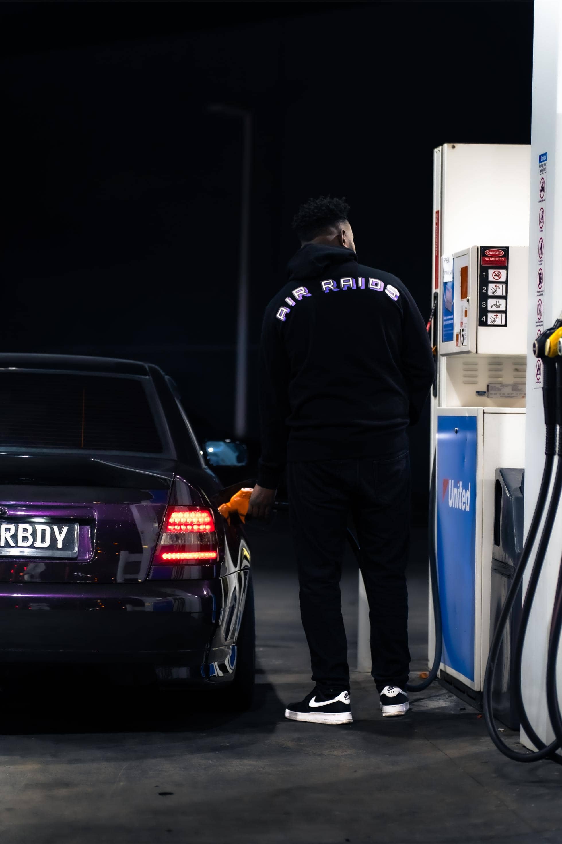 Man wearing black hoodie featuring graphic text "Air Raids" across the back, pumping petrol