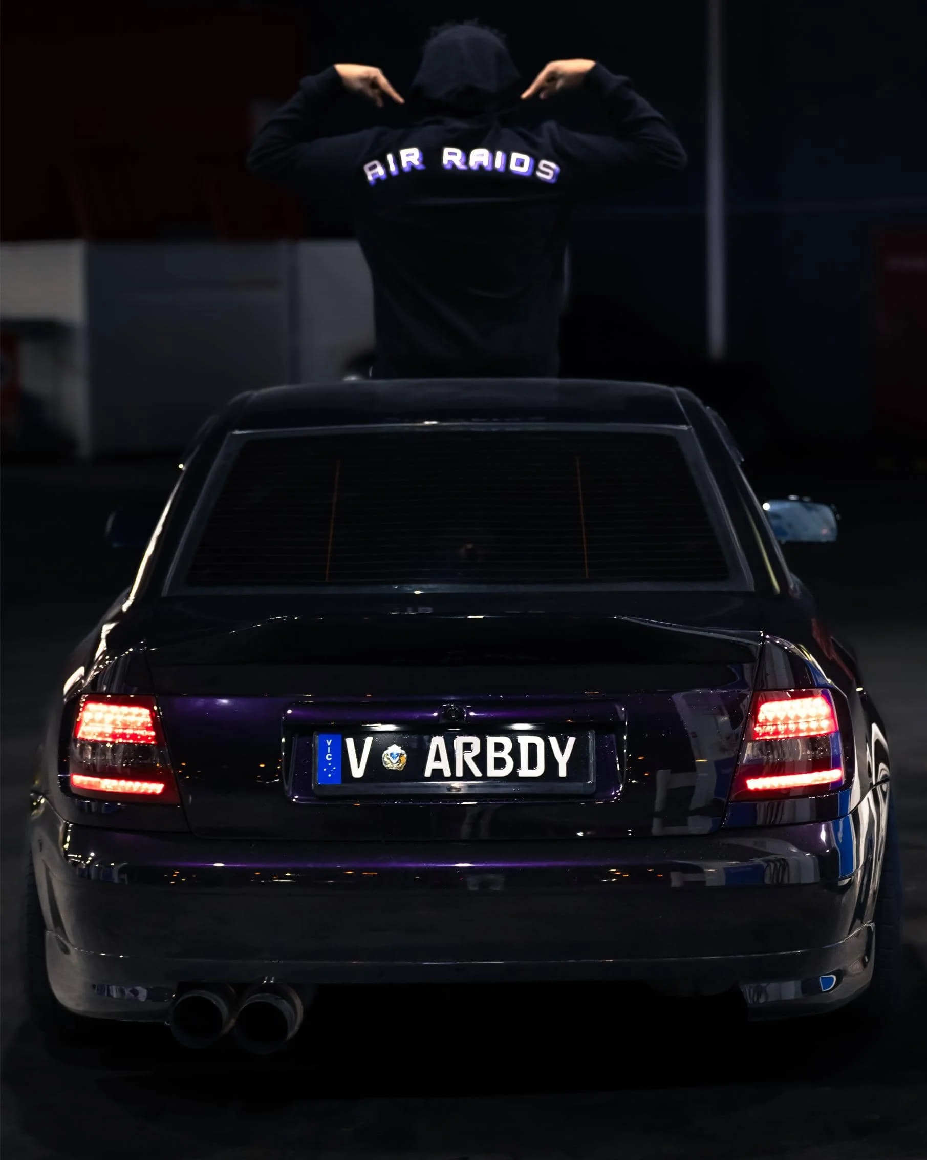 Man wearing black hoodie featuring graphic text "Air Raids" across the back, sitting on top of purple car