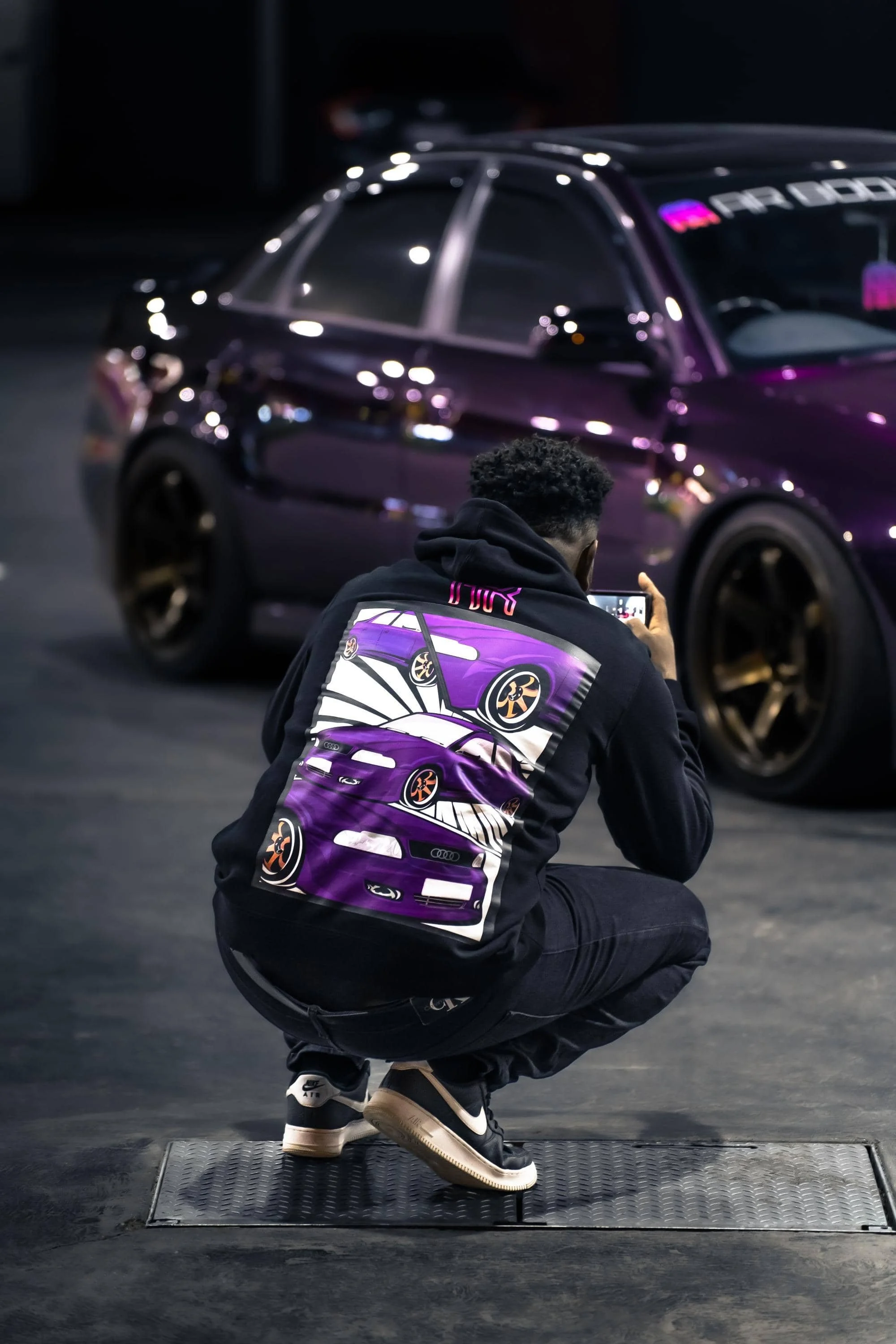 Man wearing hoodie featuring purple audi illustration and AR Bodyworks logo, kneeling in front of a blurred purple car