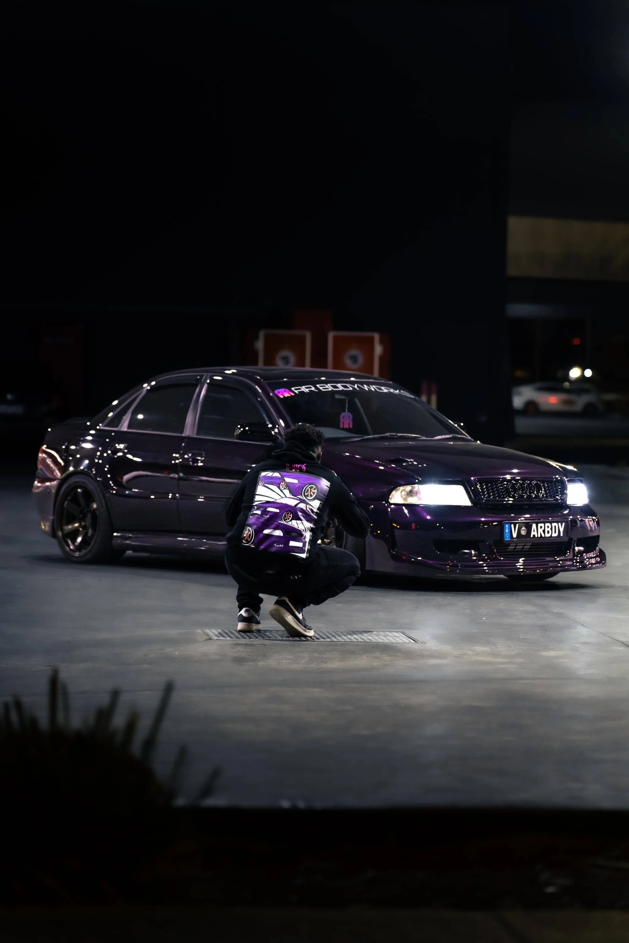 Man wearing hoodie featuring purple audi illustration and AR Bodyworks logo, kneeling in front of a blurred purple car