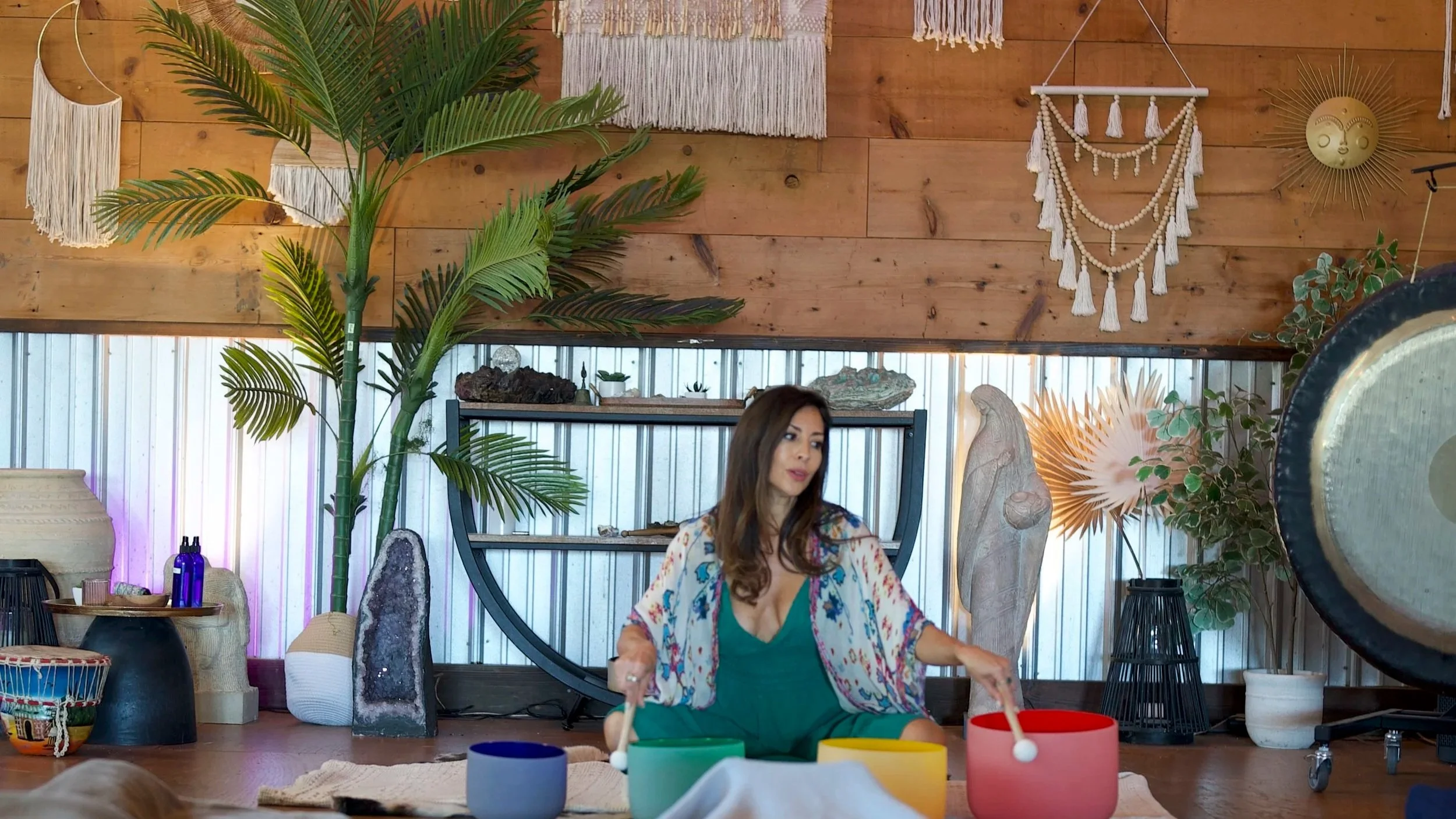 Woman sitting on the floor playing a colorful singing bowl set, surrounded by bohemian decor including large plants, driftwood, wall hangings, and a gong.